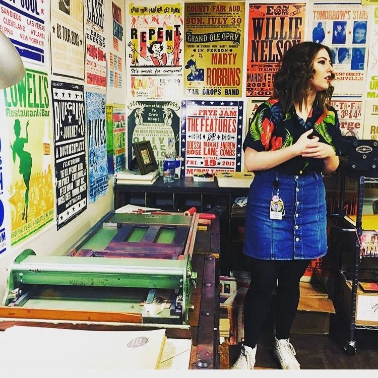 A woman standing in a room decorated with colorful posters and flyers on the wall, next to a vintage printing press.