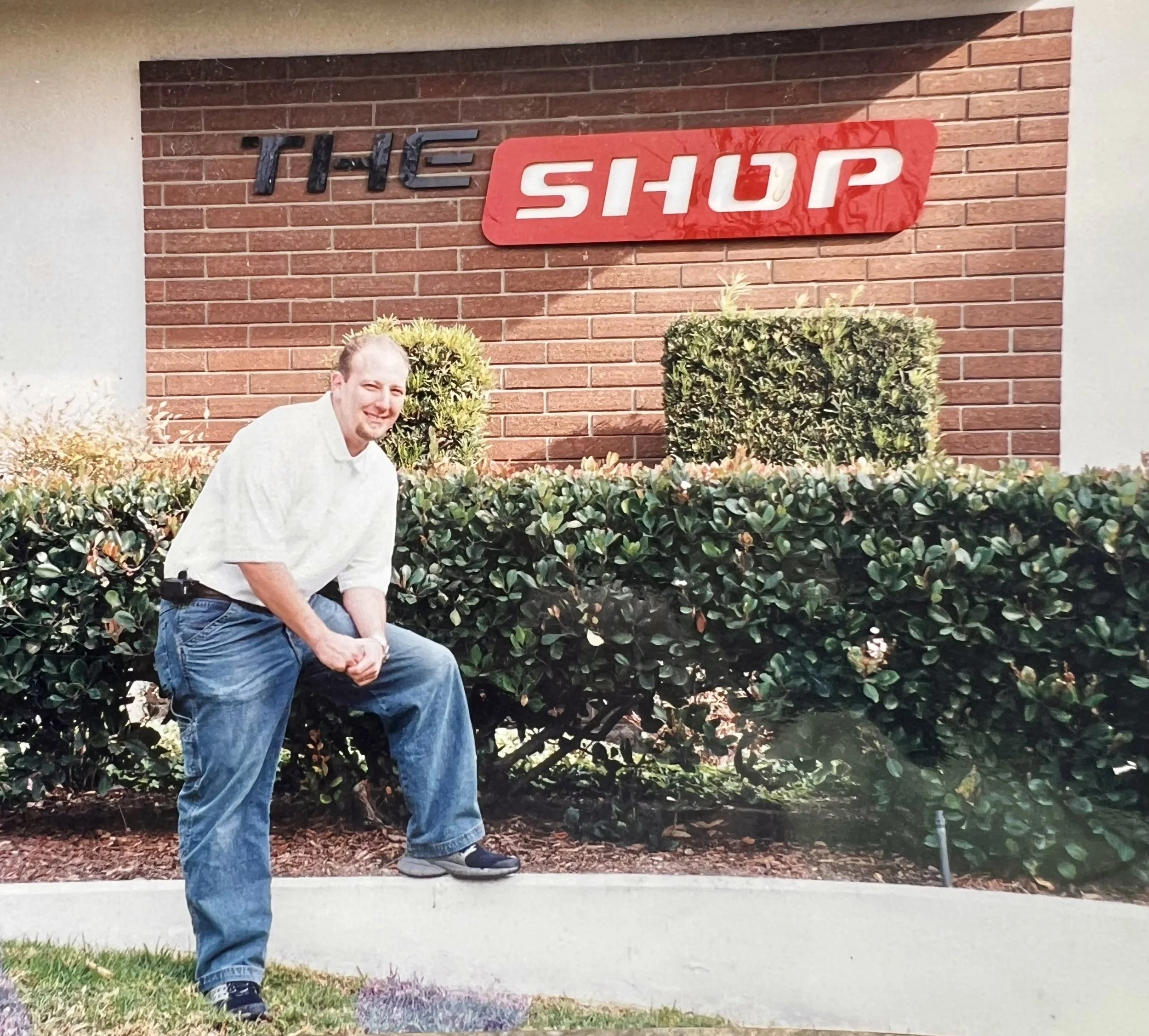 A man crouching in front of a bush and a brick wall with a red sign that says 'The Shop' in white letters.