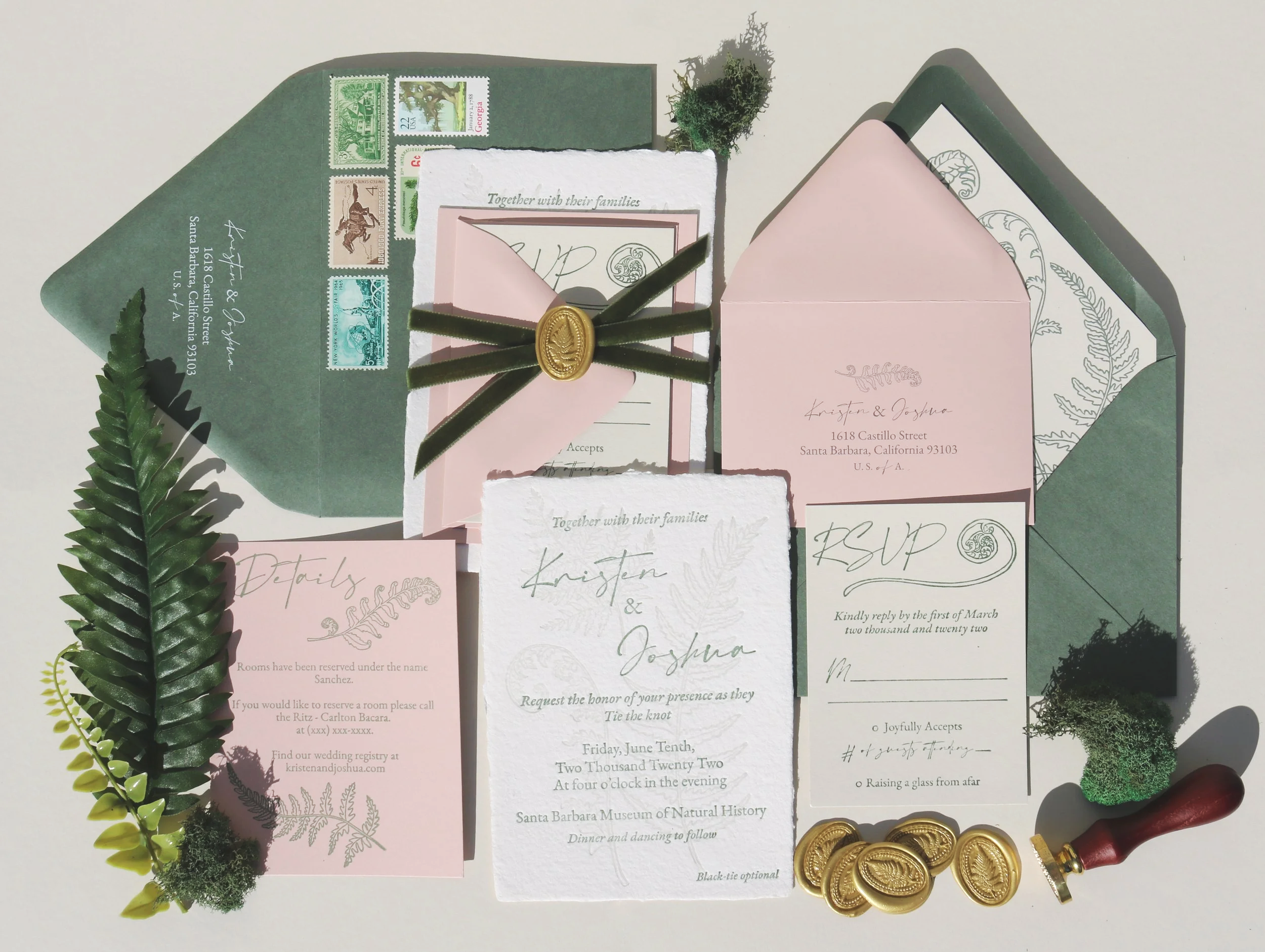 A flat lay of wedding invitation stationery with greenery, stamps, gold seals, and a wax stamp on a white background.