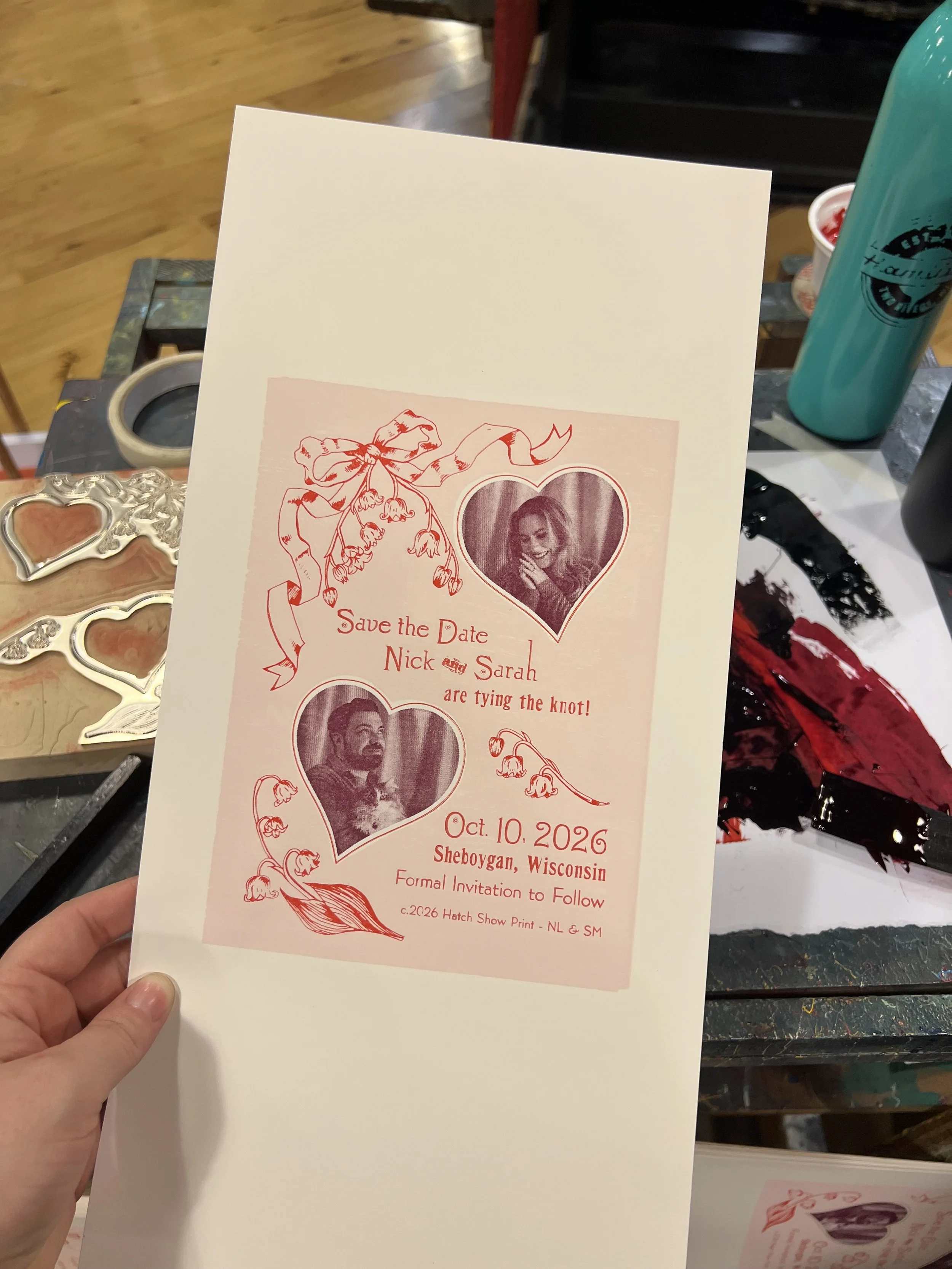 Partial view of a wedding save the date card with red floral design, heart-shaped photo frames, and text indicating the wedding date of October 10, 2026, with names Nick and Sarah, in Sheboygan, Wisconsin, held on a decorated table.