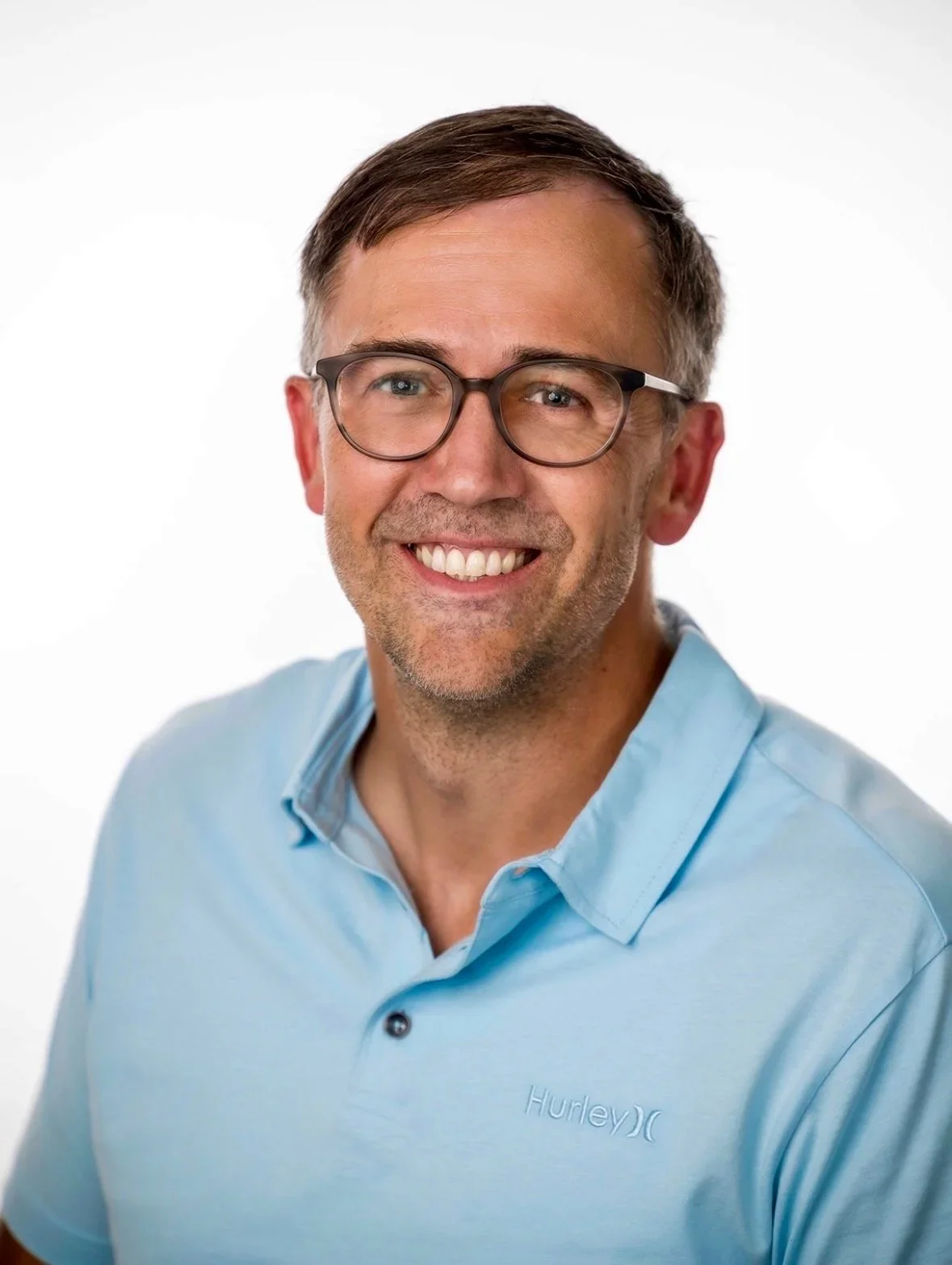 A smiling man wearing glasses and a light blue Hurley polo shirt, posing against a plain white background.