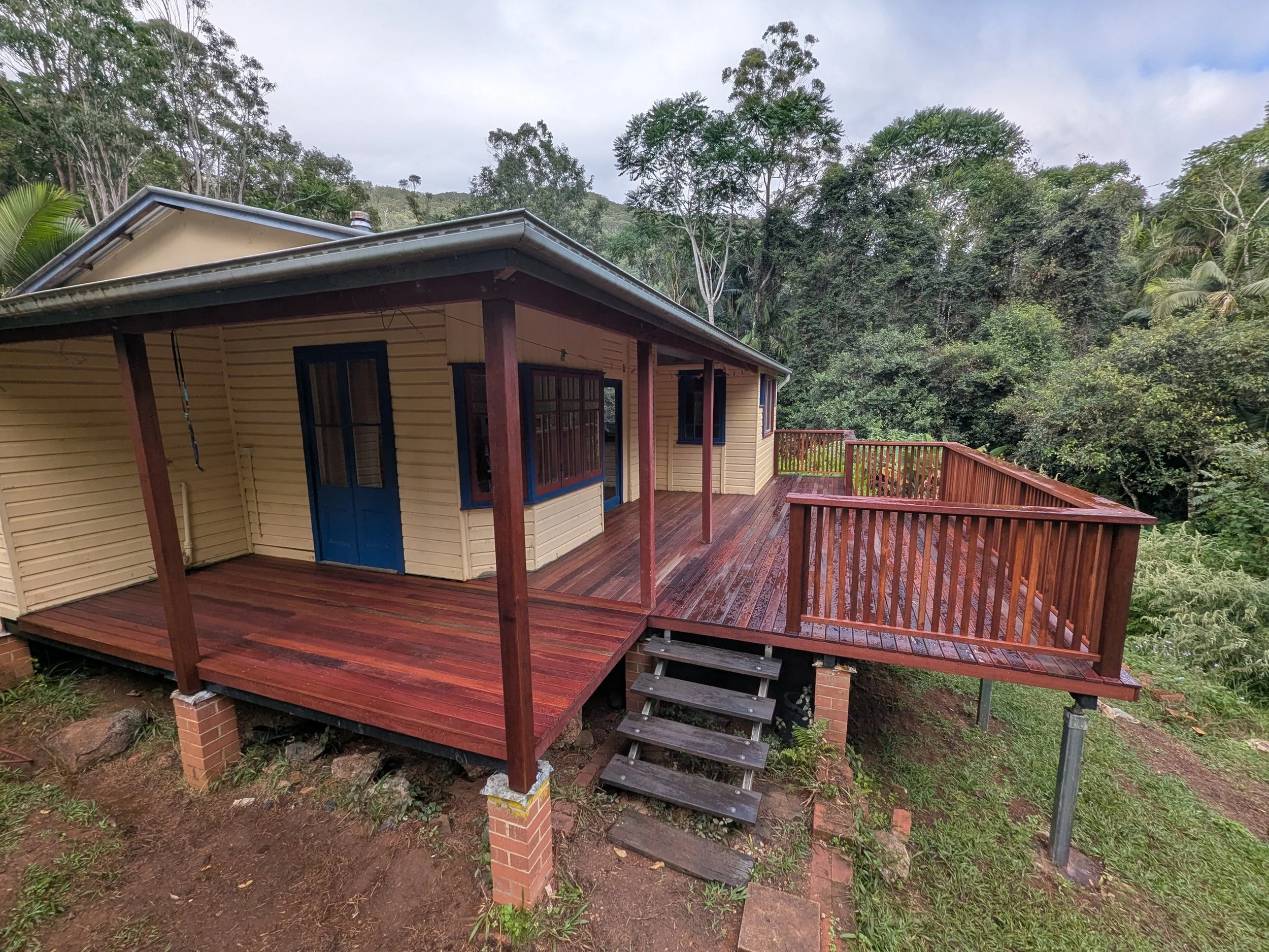 A house with a wooden deck and railing, surrounded by tall trees and lush greenery in a forested area under a cloudy sky.