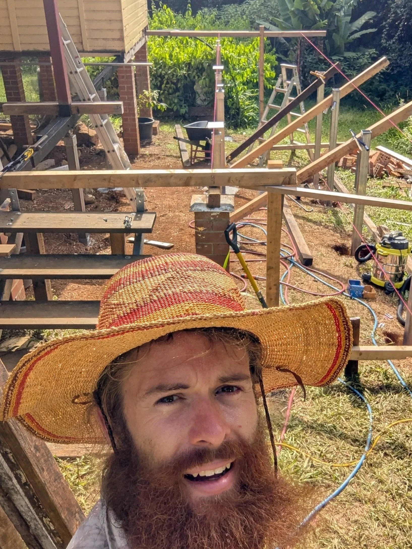 A man with a large grey beard and a straw hat smiling at the camera, while construction work is happening in the background with wooden beams, ladders, and tools.