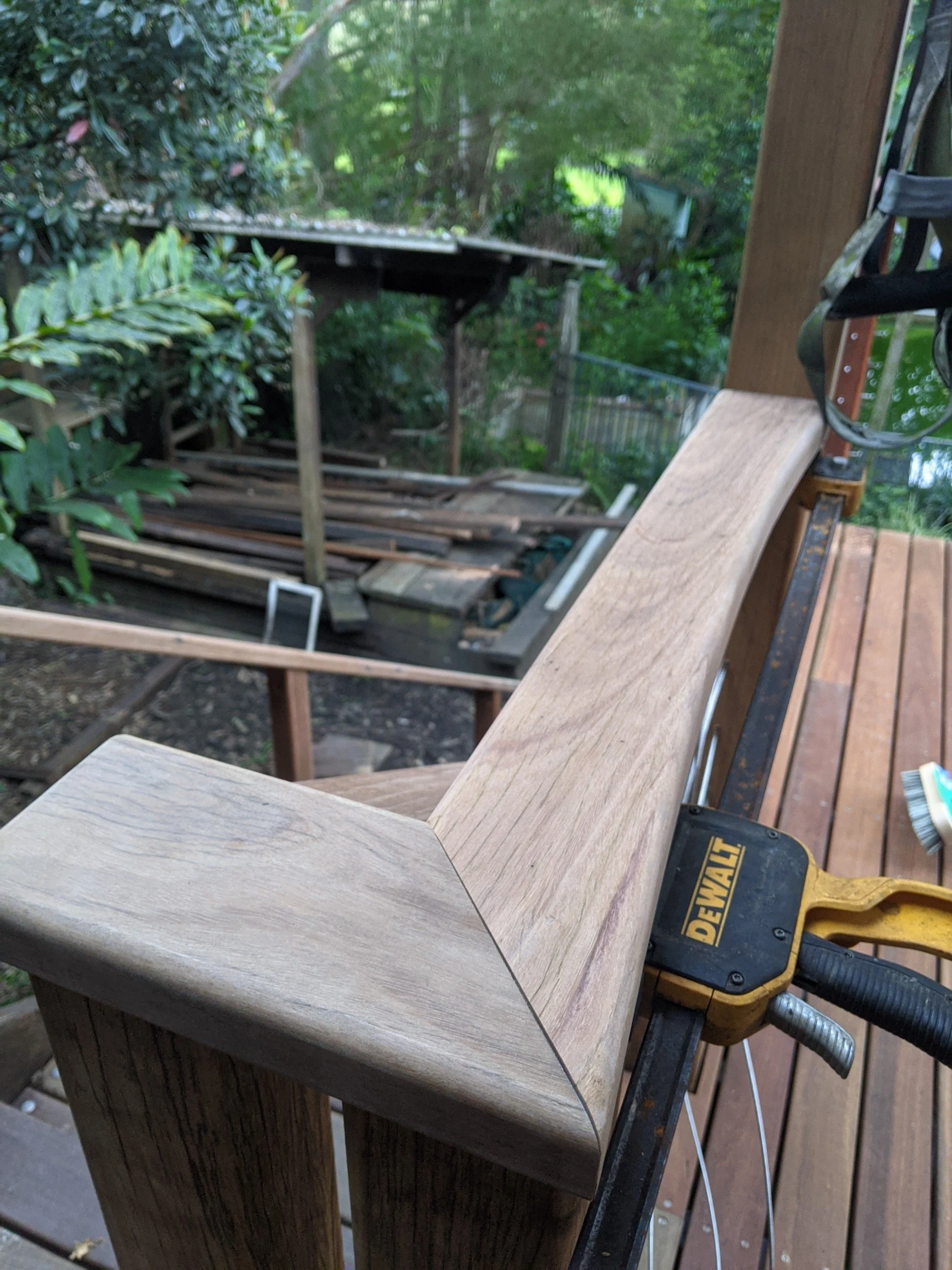 Close-up of a wooden deck railing or stair banister under construction, with a power drill visible on the right side and green foliage in the background.