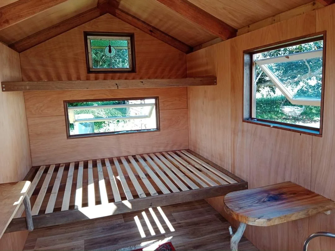 Interior of a small wooden room with two windows, one open and one closed, a wooden bed frame without a mattress, and a small wooden table or shelf in the corner.
