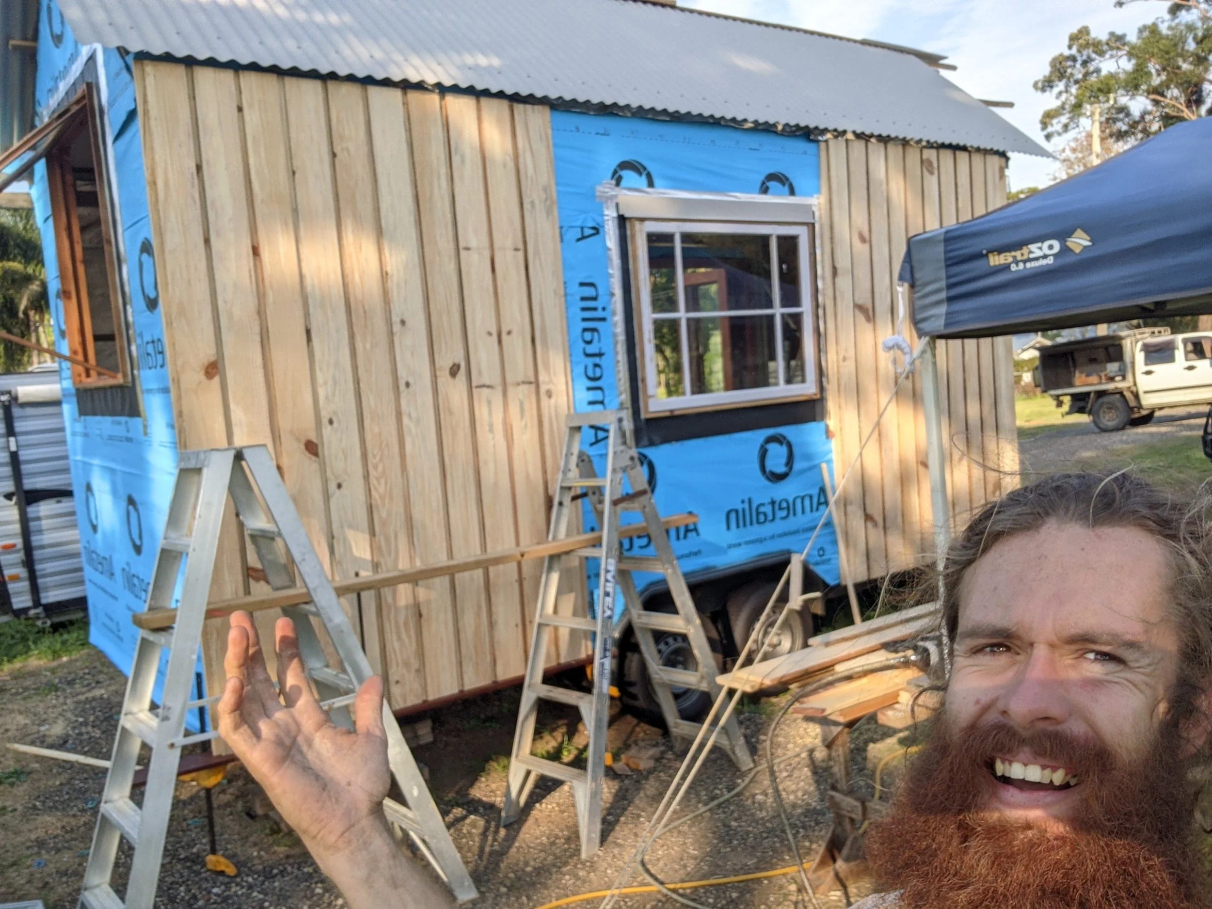 A person with red hair and a beard is smiling brightly while taking a selfie outdoors. In the background, there is a wooden structure under construction, with visible wood panels and a window frame. A ladder and construction materials are also visibl