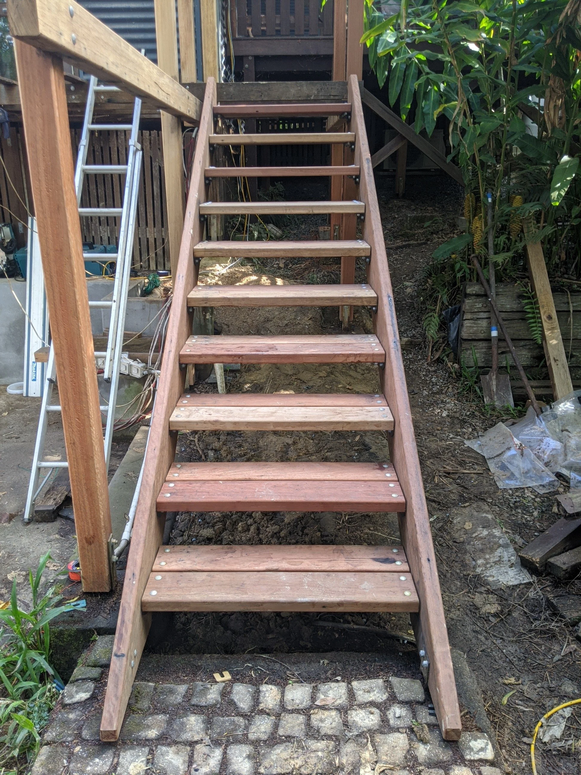 Wooden outdoor staircase under construction with visible steps and side rails, surrounded by garden and construction tools.