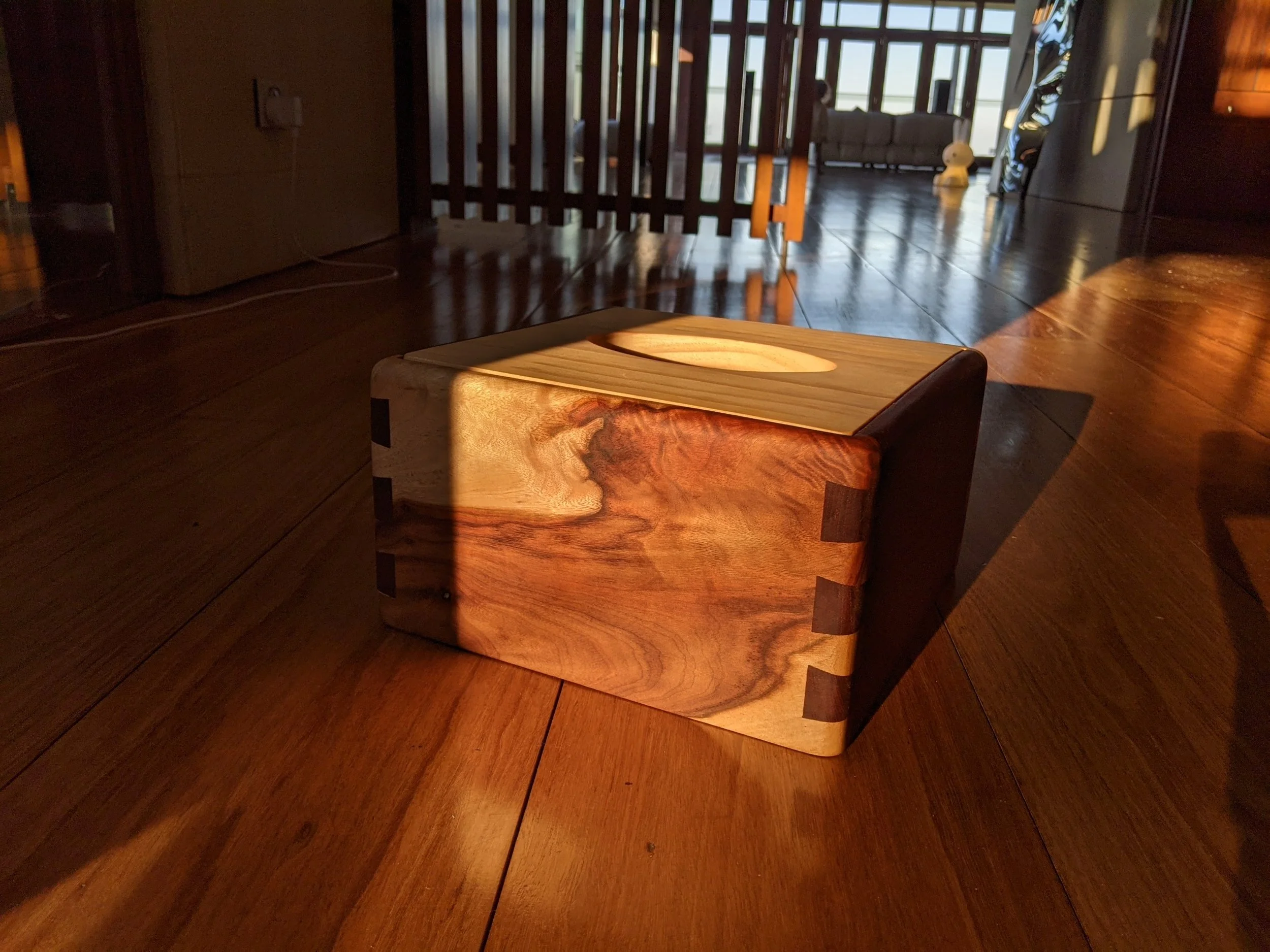 A wooden cube-shaped side table with a circular hollow in the top surface, placed on a wooden floor near a dark wood wall, with sunlight casting shadows.