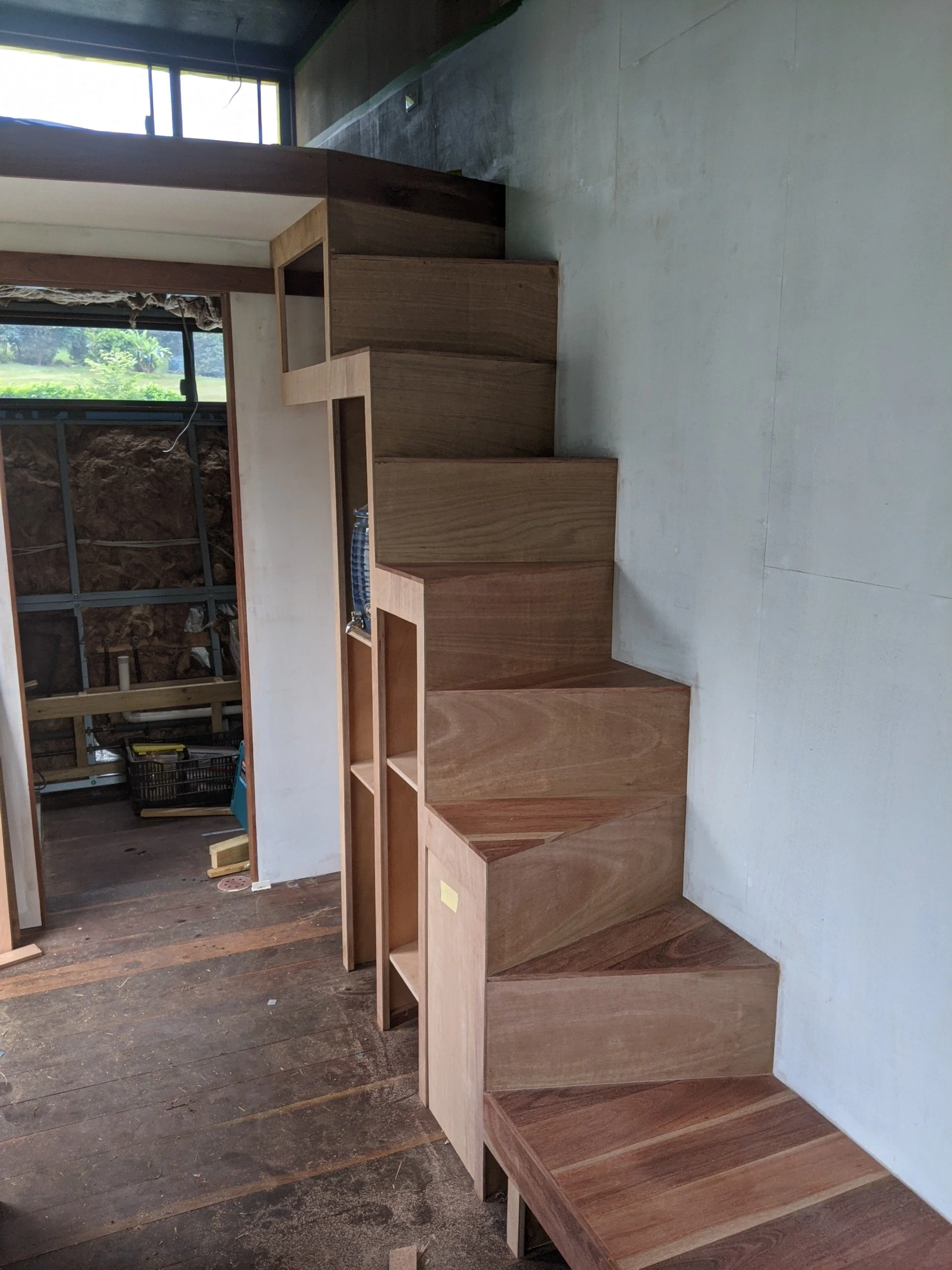 Wooden staircase in a room under construction, with a window showing green outdoor scenery and an unfinished wall.