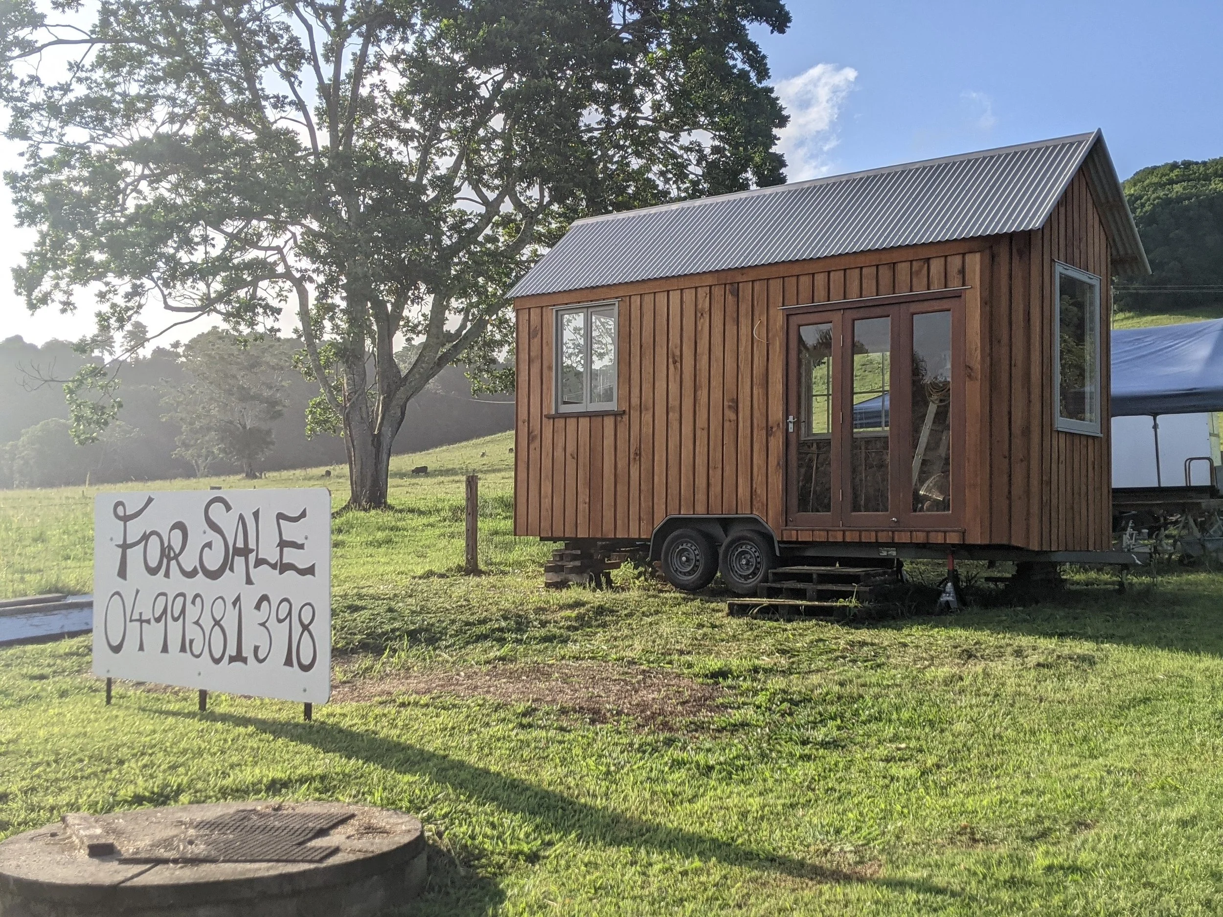 A small wooden house on wheels with a metal roof, situated on green grass beside a tree. A sign in front of the house reads 'For Sale' with a phone number.