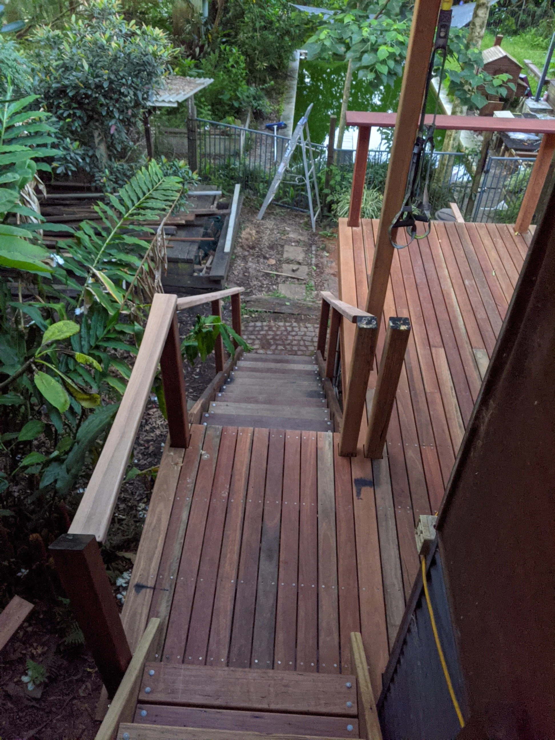 A wooden deck with stairs leading down to a dirt path, surrounded by lush green plants and trees near a body of water.