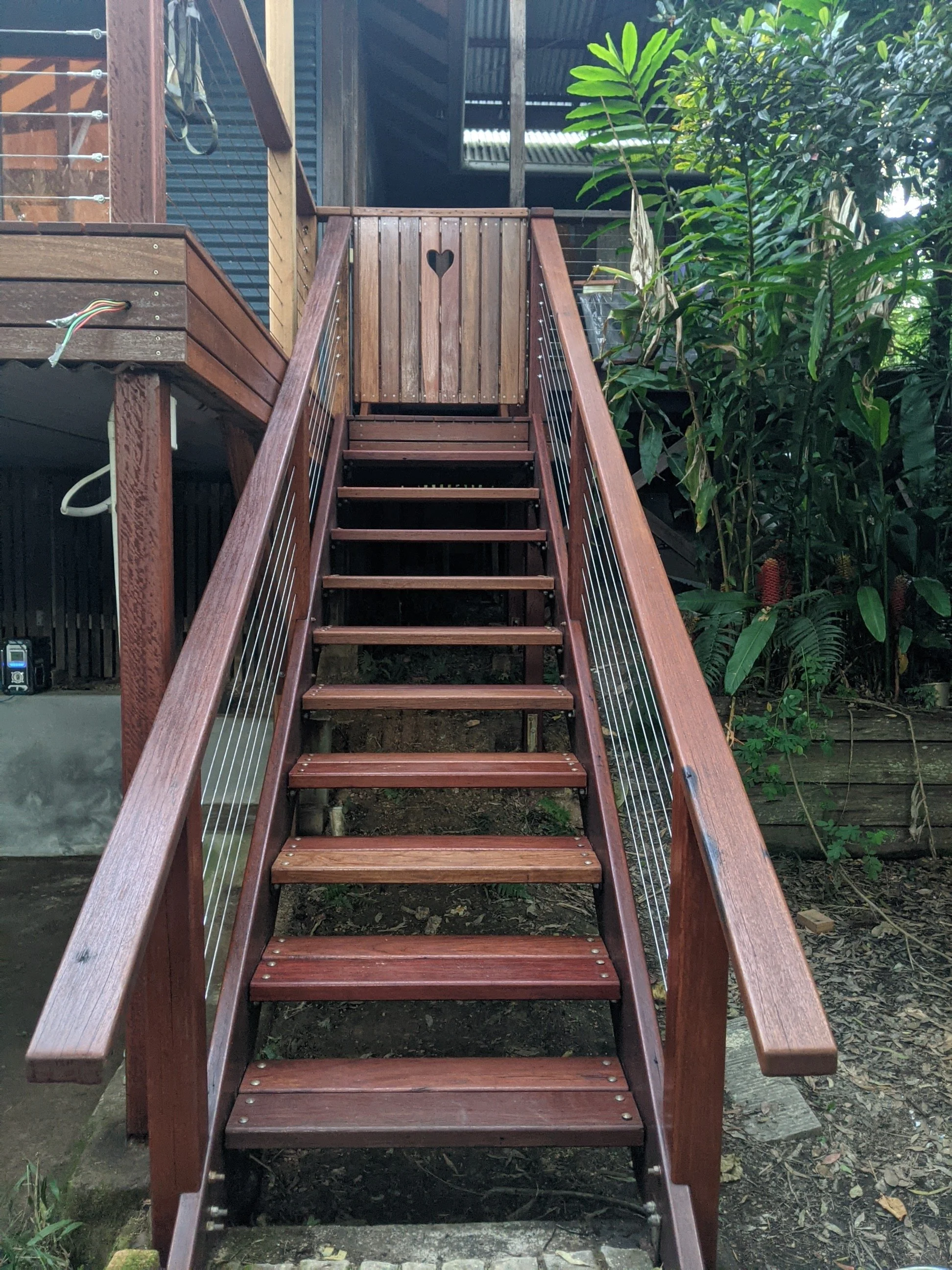 Wooden outdoor staircase with handrails, leading up to a small deck with a wooden gate that has a heart-shaped cutout. Green plants and trees surround the staircase.
