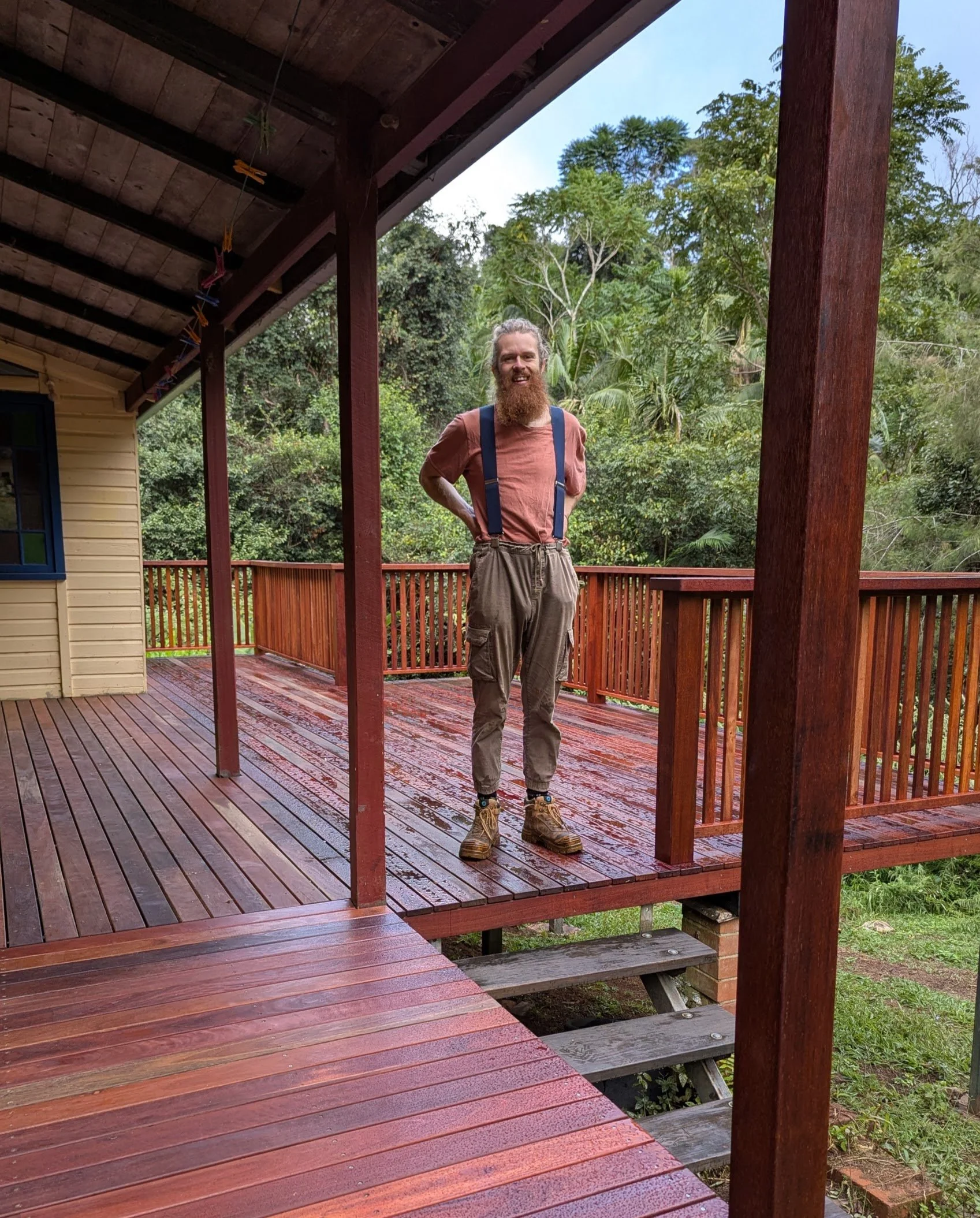 A man with long hair and a beard standing on a wooden deck outdoors, wearing a brown shirt, suspenders, baggy pants, and hiking boots, smiling at the camera with a lush green background.