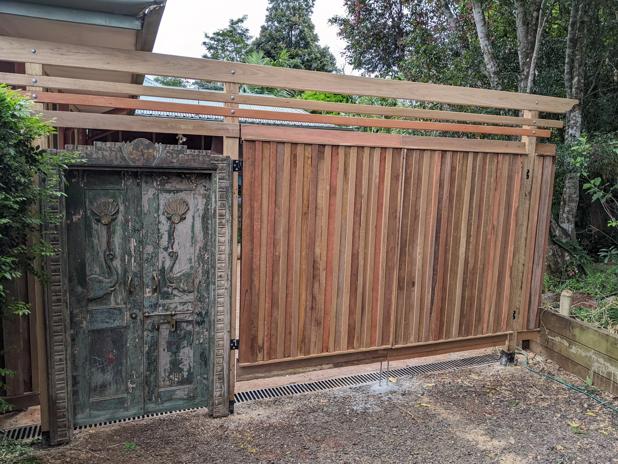A wooden fence under construction with vertical and horizontal wooden planks, surrounding a backyard area with trees and bushes, and an old weathered decorative metal gate to the left.
