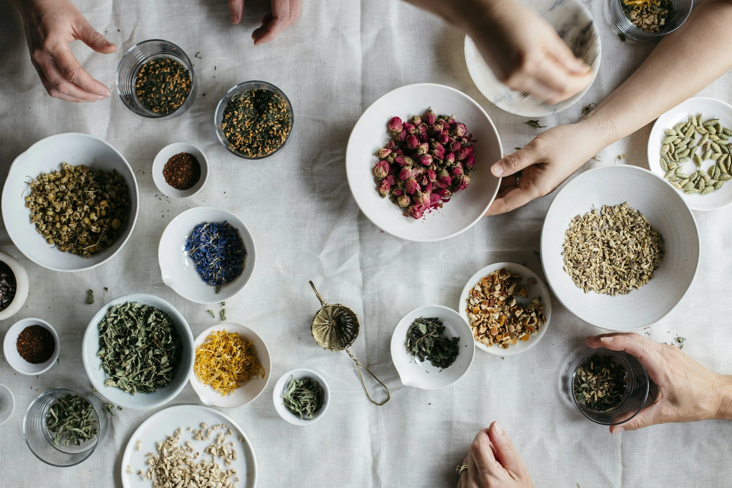 Top-down view of various dried herbs and flowers in bowls, with hands handling some of the bowls on a white tablecloth.