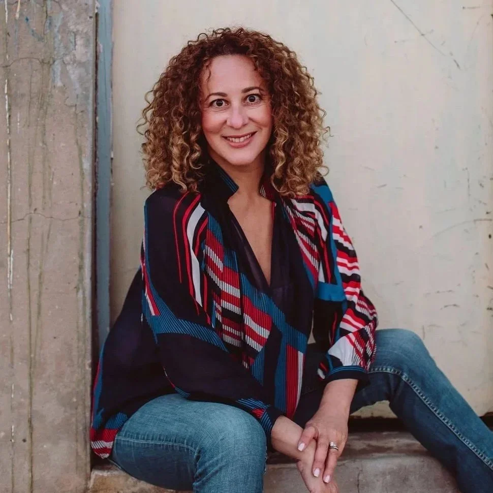 A woman with curly hair sitting on the ground near a wall, smiling at the camera. She wears a colorful, geometric patterned jacket and blue jeans.
