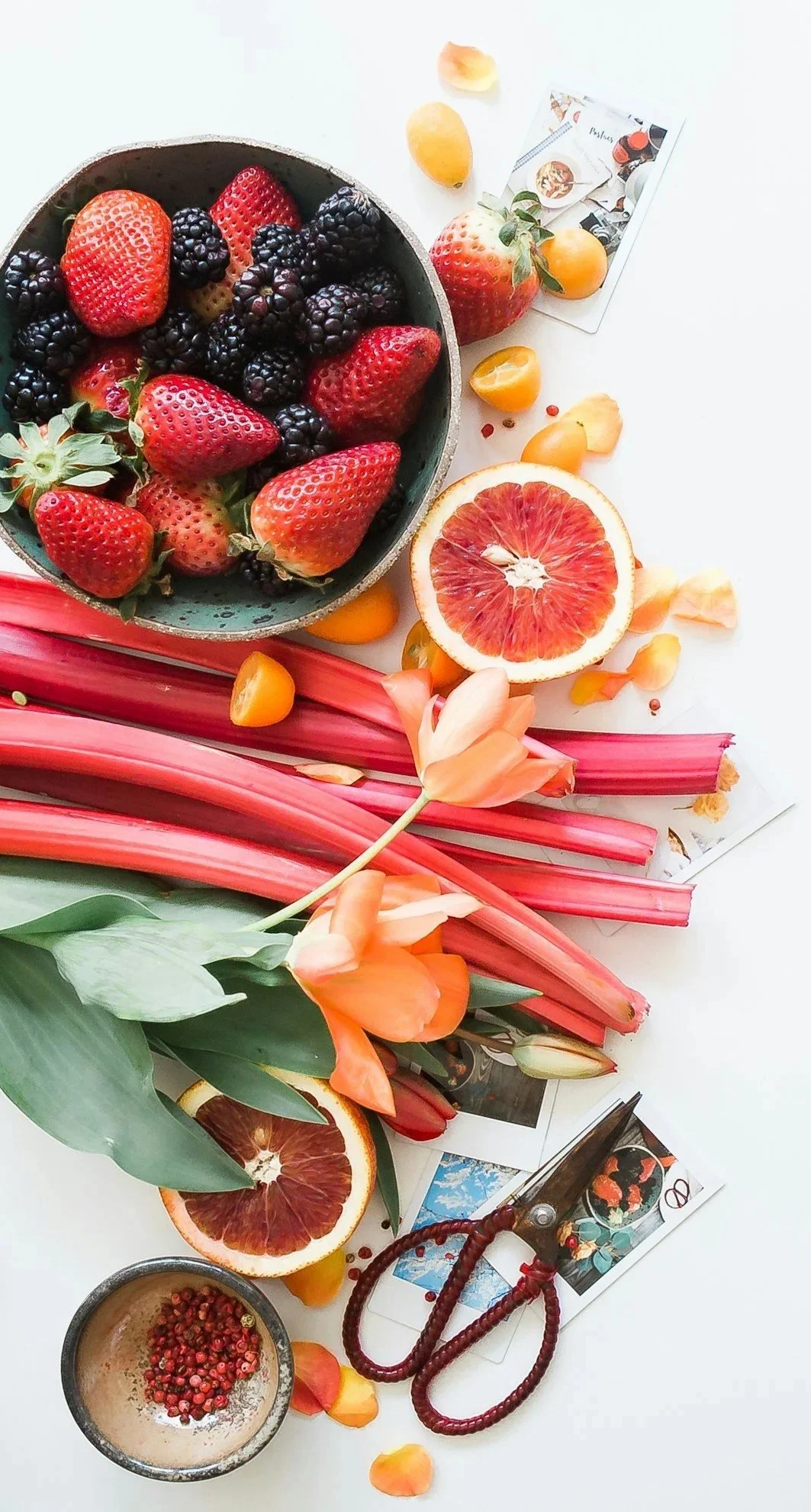 A bowl of fresh strawberries, blackberries, and a halved grapefruit, with flower petals and assorted flowers, including lilies, on a white surface.