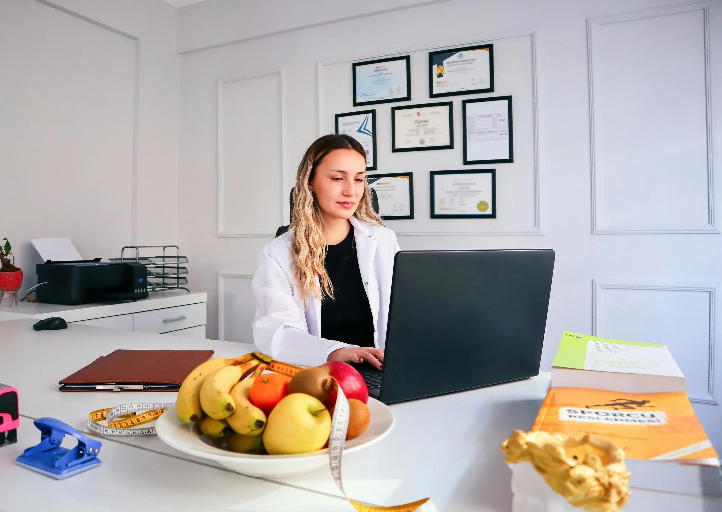A woman in a white coat sitting at a desk with a laptop in a bright office, with framed certificates on the wall behind her. The desk has a fruit bowl filled with bananas, apples, and oranges, a measuring tape, a book, and some stationery.