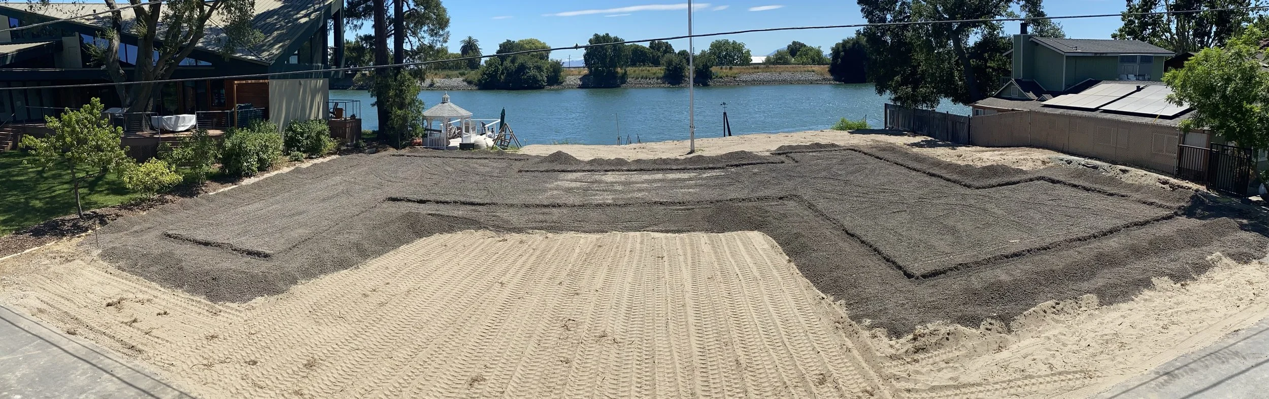 A construction site with sand and gravel, partially leveled, in a backyard near water with trees and houses in the background.