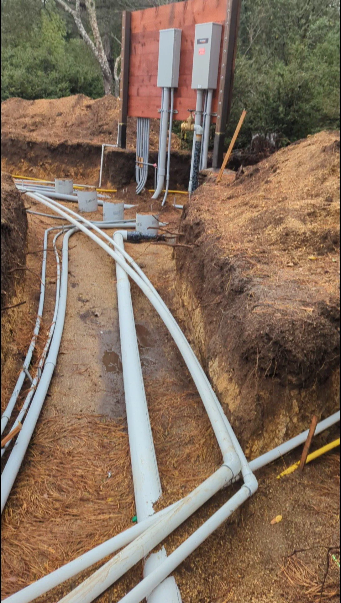 Underground utility installation with multiple white pipes and electrical boxes on a construction site surrounded by dirt and trees.