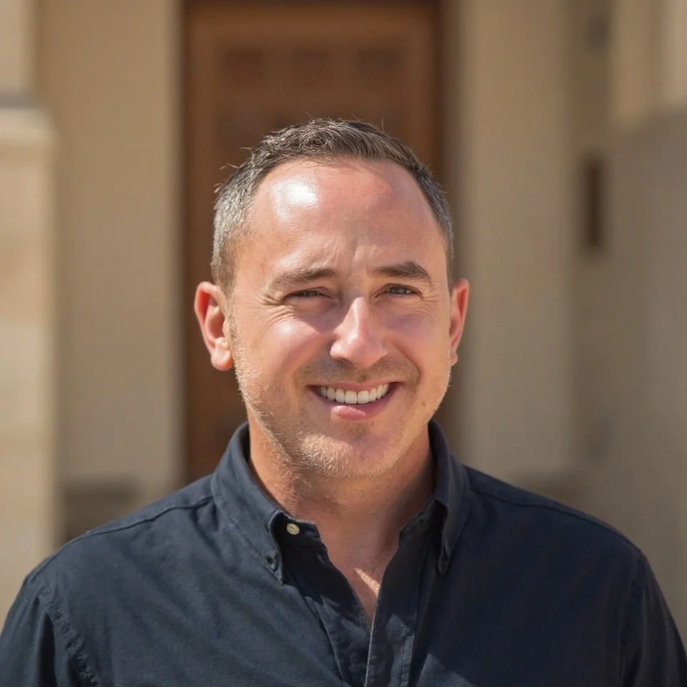 Portrait of a man smiling outdoors, wearing a black button-up shirt with a blurred building background.
