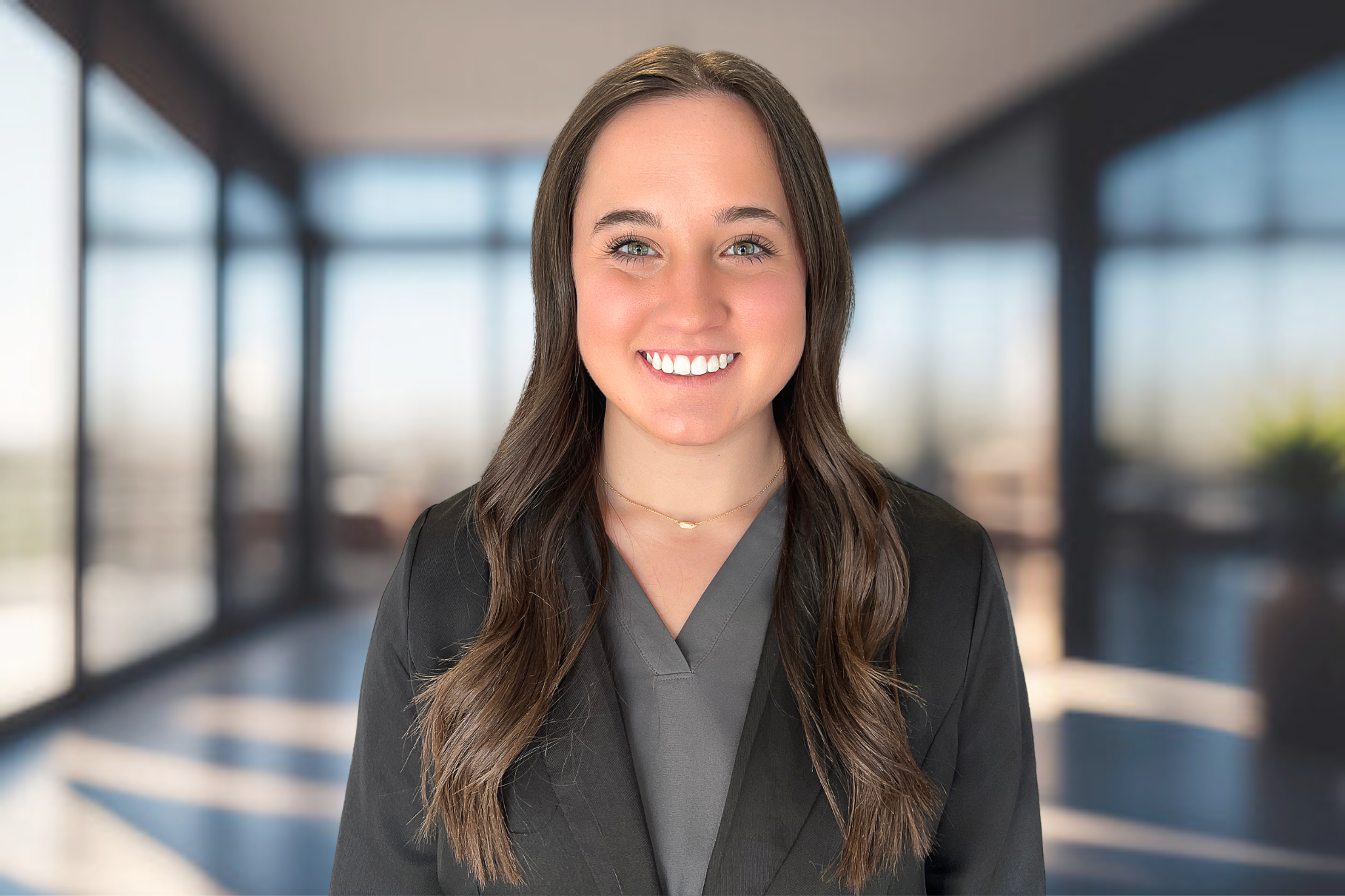 A young woman with long brown hair, wearing a dark blazer and a gray top, smiling in an office corridor with large windows and blurred background.