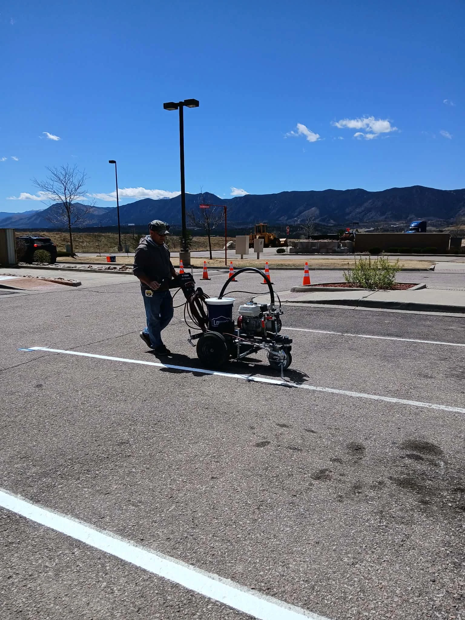 A man operating equipment in a parking lot under a clear blue sky with distant mountains in the background.