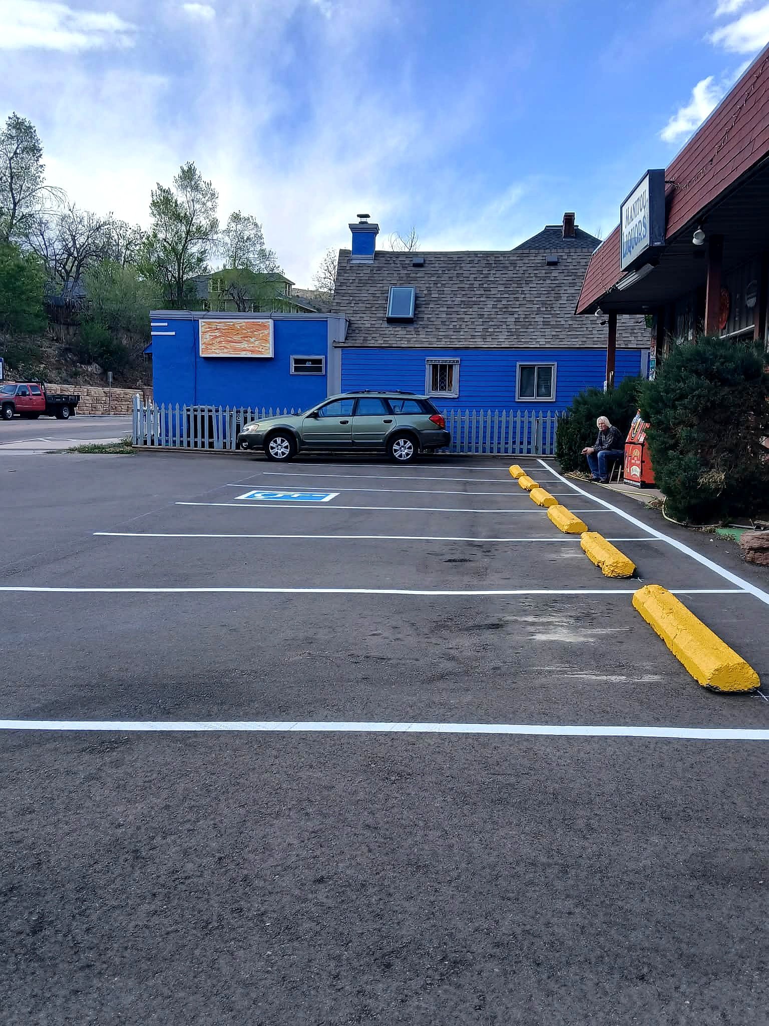 Empty parking lot with a disabled parking space marked in blue and yellow concrete parking stops; a man sitting on a bench near a bush and a building with a sign, cars and a blue building with a chimney against a partly cloudy sky.
