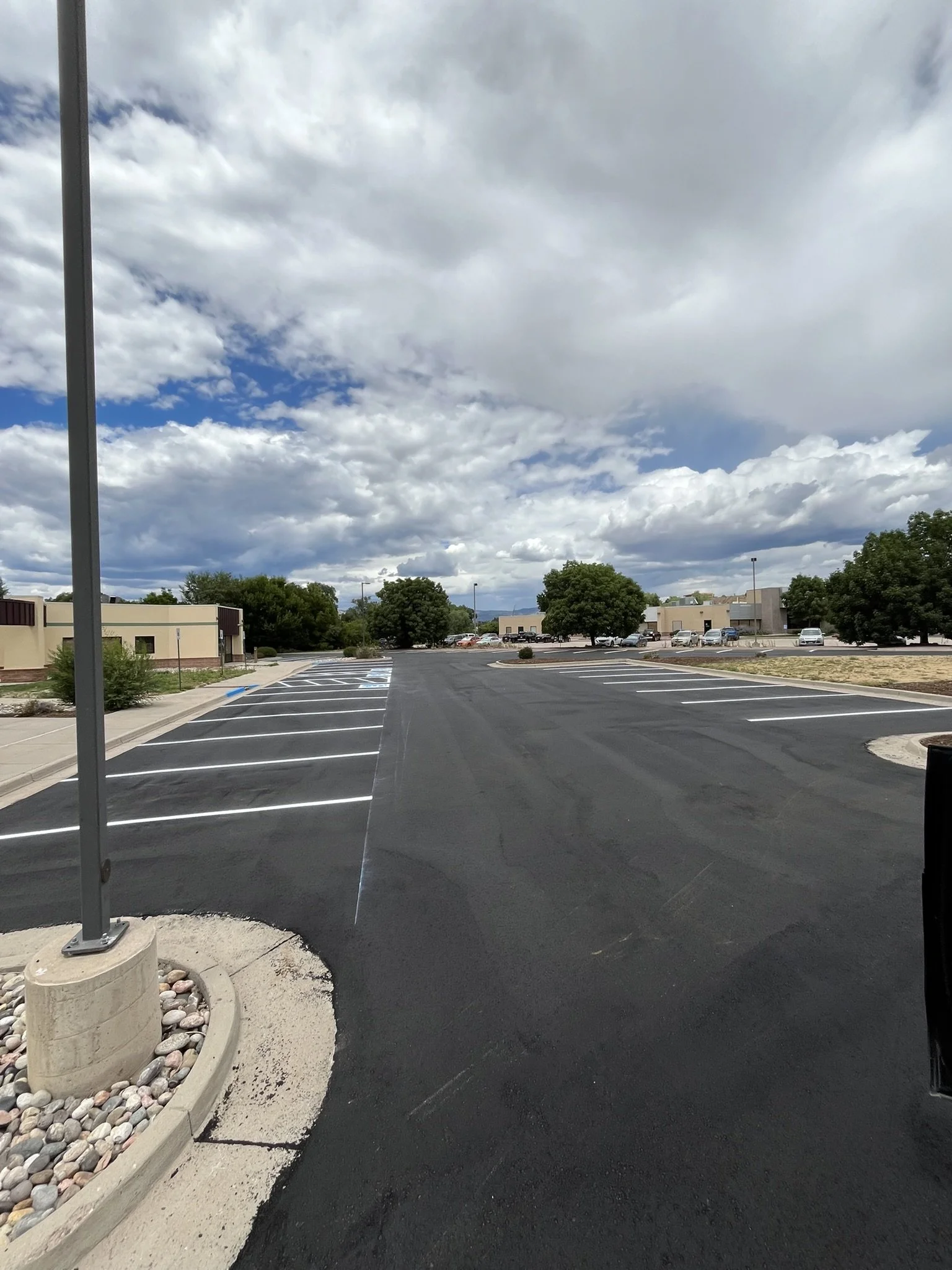 Empty parking lot with freshly paved asphalt, marked parking spaces, and some trees in the background under a partly cloudy sky.