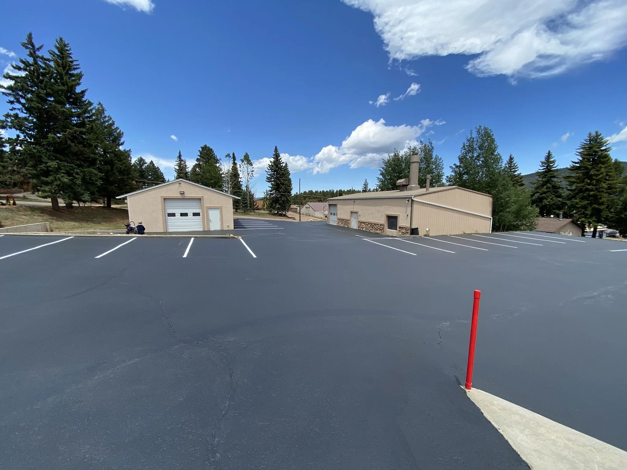Empty parking lot surrounded by trees, buildings, and a mountain with blue sky and clouds overhead.