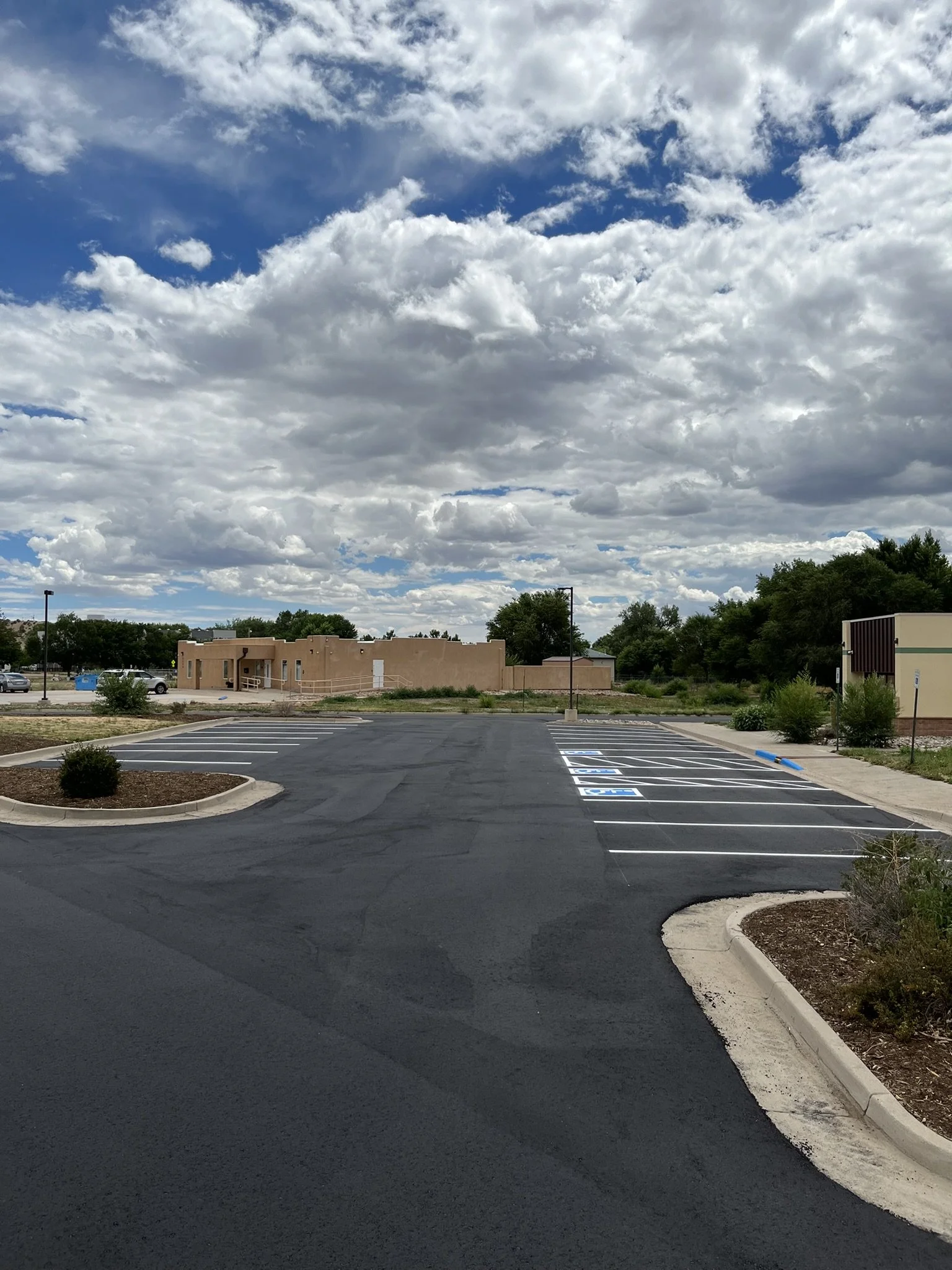 Empty parking lot with parking spaces marked, a building in the background, and a partly cloudy sky.