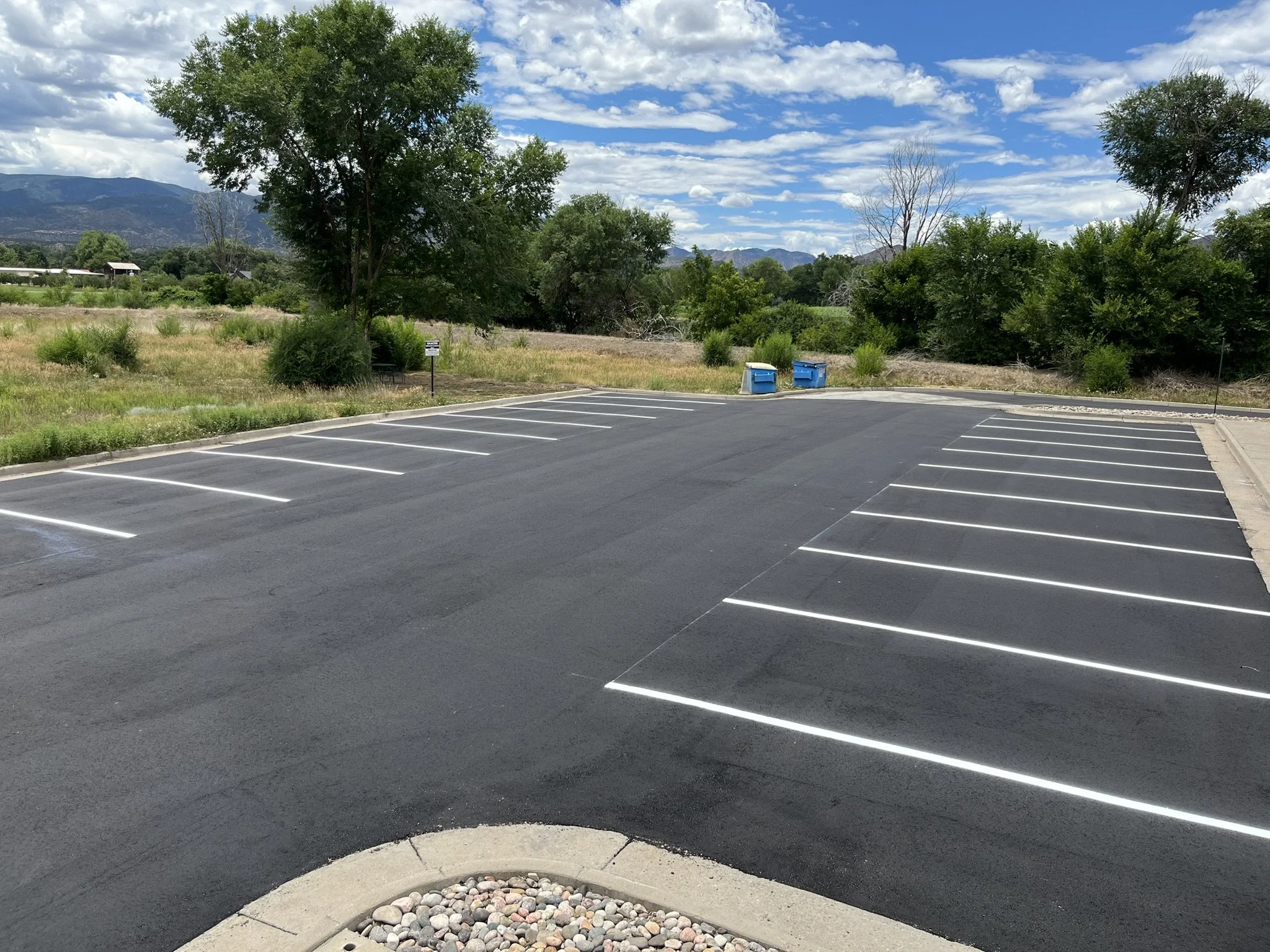An empty parking lot with empty parking spaces, a few trees, a grassy area, blue recycling bins, and a mountain range in the background under a partly cloudy sky.