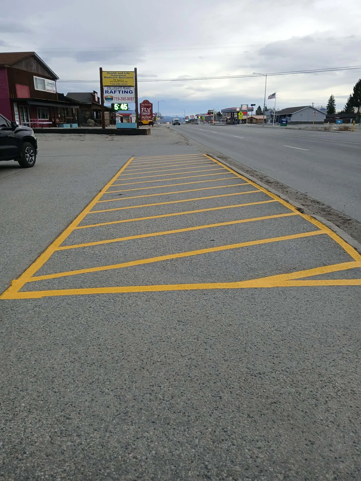 Empty parking lot with yellow striped parking spaces in a small town with buildings, hills, and street signs visible in the background.