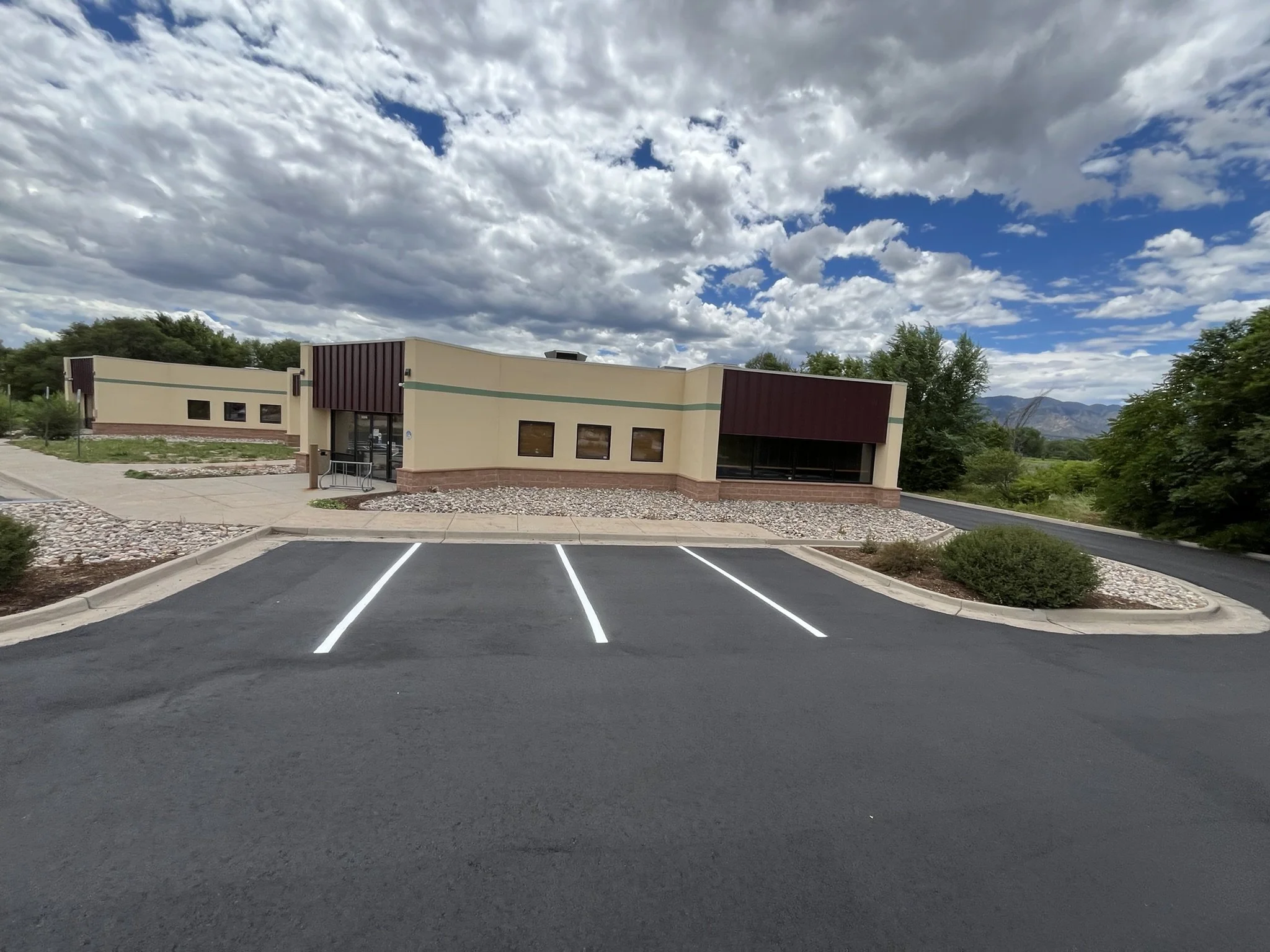 Empty parking lot in front of a beige building with a brown accent on the roof, surrounded by trees and mountains under a partly cloudy sky.