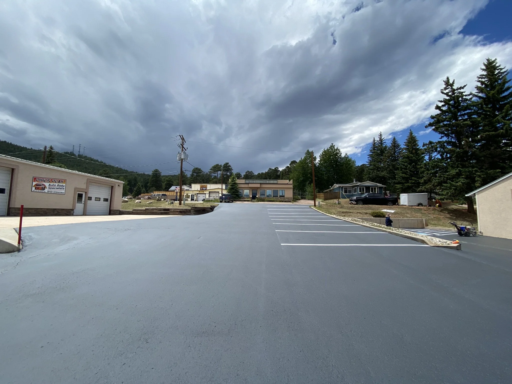 Empty parking lot with a building labeled Pikes Peak Auto Body Specialists, surrounded by trees and hills, under a cloudy sky.