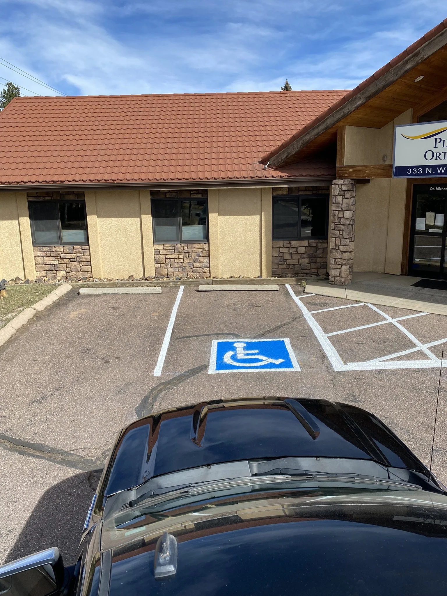 View of a parking lot with a handicap parking space marked with a blue sign and a building with a stone and beige exterior, a red-tiled roof, and windows, with the front of a black vehicle visible in the foreground.