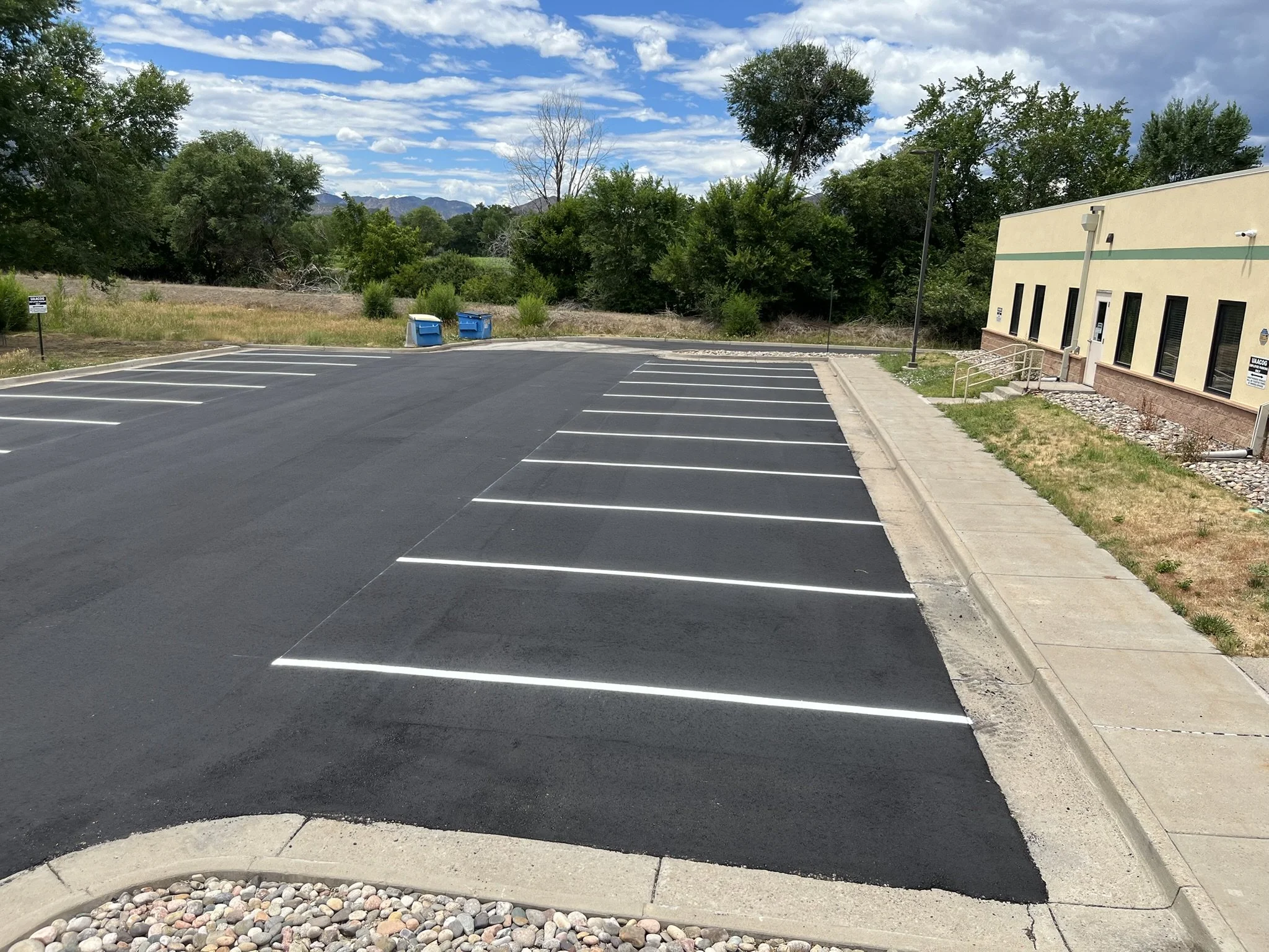 Empty parking lot with marked spaces, two blue trash bins, trees and a building with a beige exterior on a partly cloudy day.
