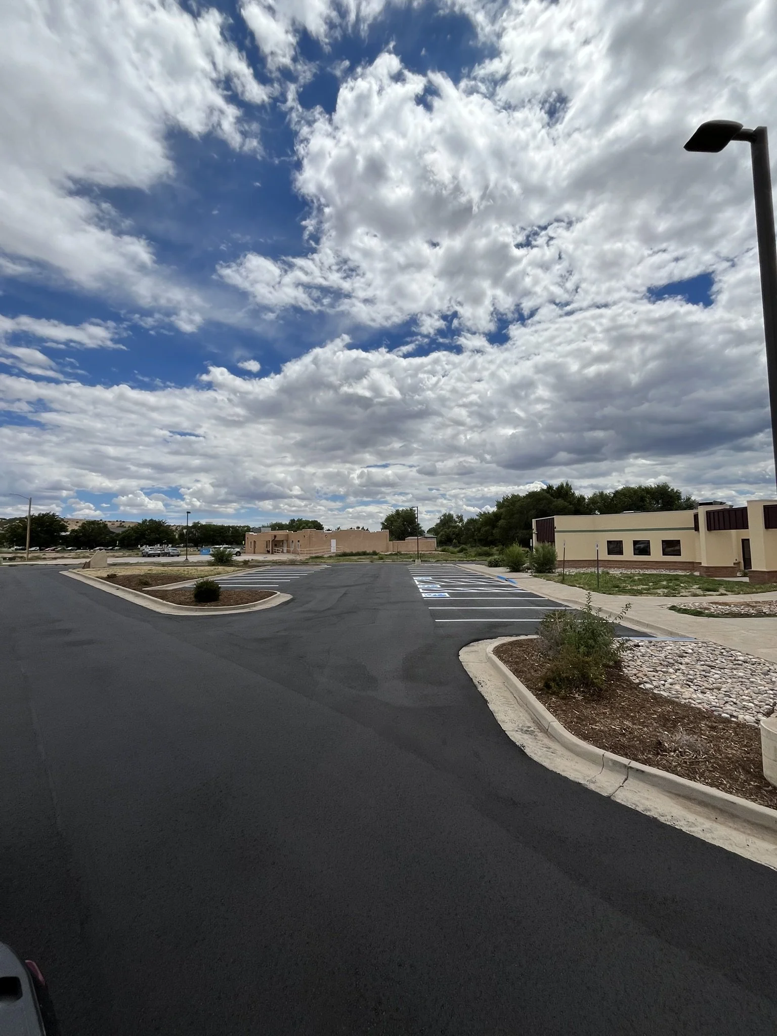 Empty parking lot with marked spaces, small landscaped areas, single-story commercial buildings, trees in the background, partly cloudy sky.