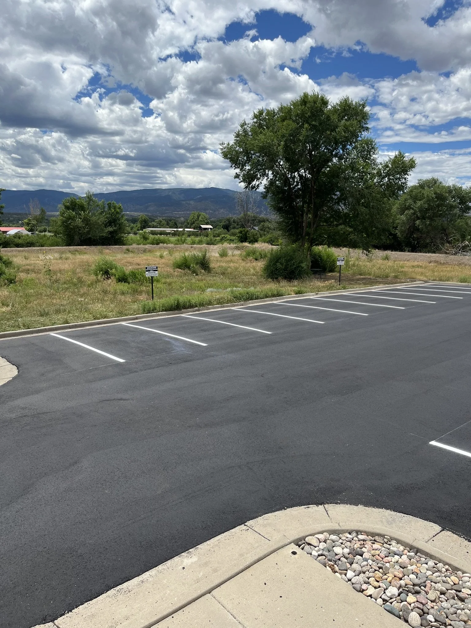 Empty parking lot with painted white lines, a tree, a small patch of grass, and a round concrete planter filled with rocks. In the background, there are green fields, trees, and distant mountains under a partly cloudy sky.