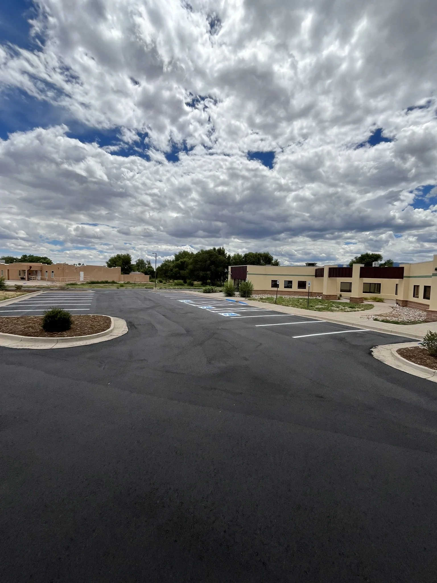 Empty parking lot with marked parking spaces and surrounding landscaping under a partly cloudy sky.
