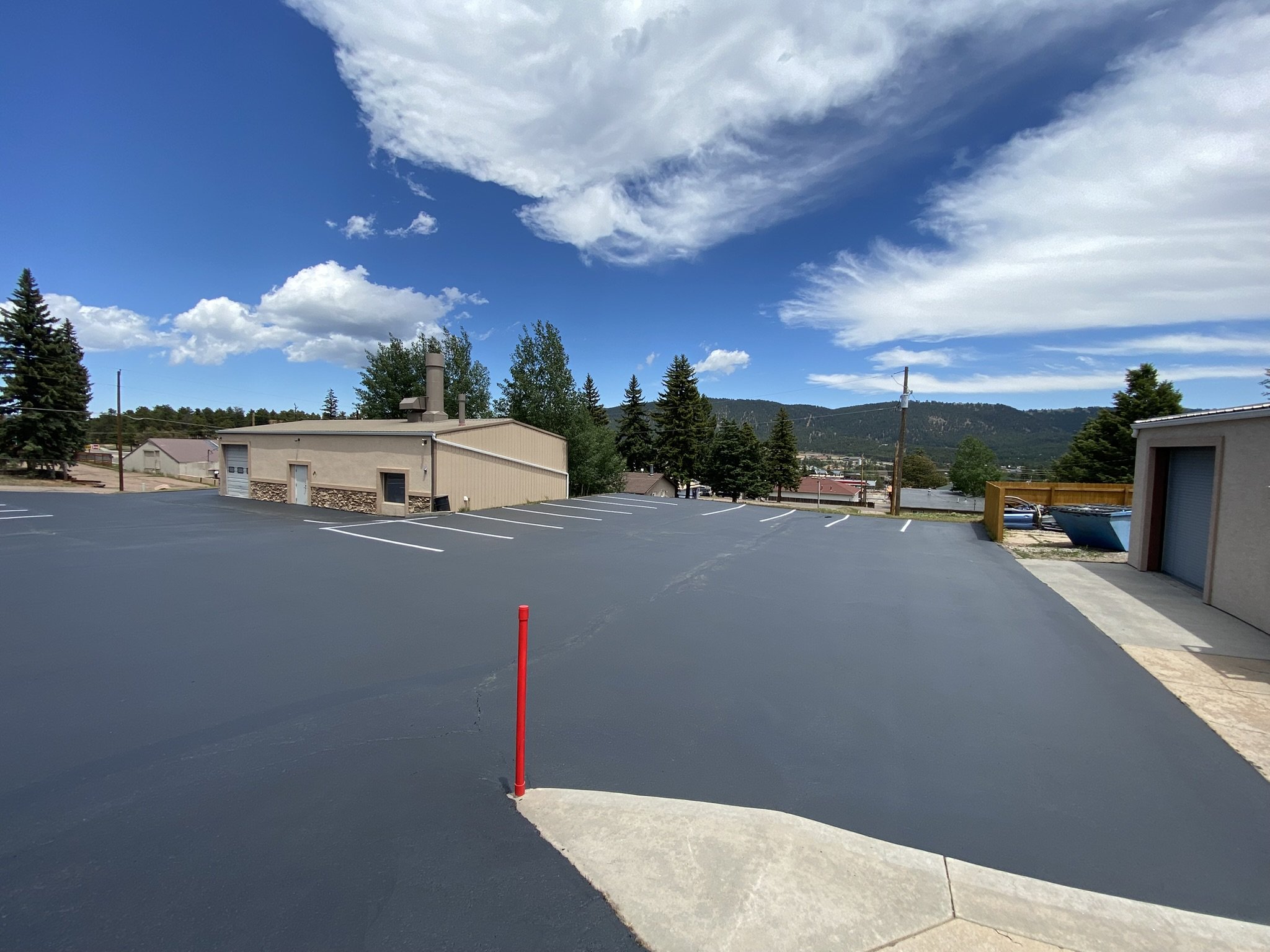 Empty parking lot with fresh asphalt, a red marker post, buildings, trees, and mountains in the background under a partly cloudy sky.