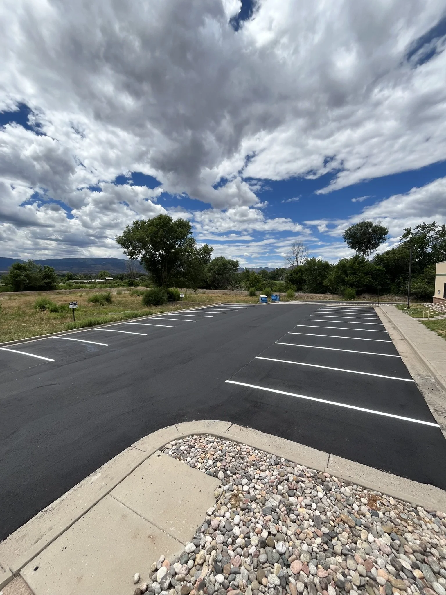 Empty parking lot with marked parking spaces, trees and mountains in the background, under a partly cloudy sky.