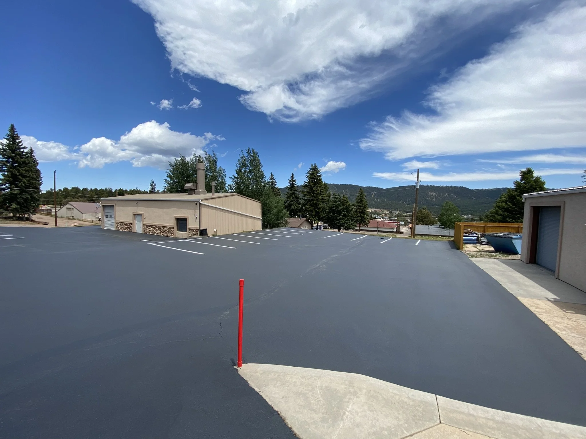 Empty parking lot with a red pole marker, buildings, trees, and mountains in the background under a partly cloudy sky.