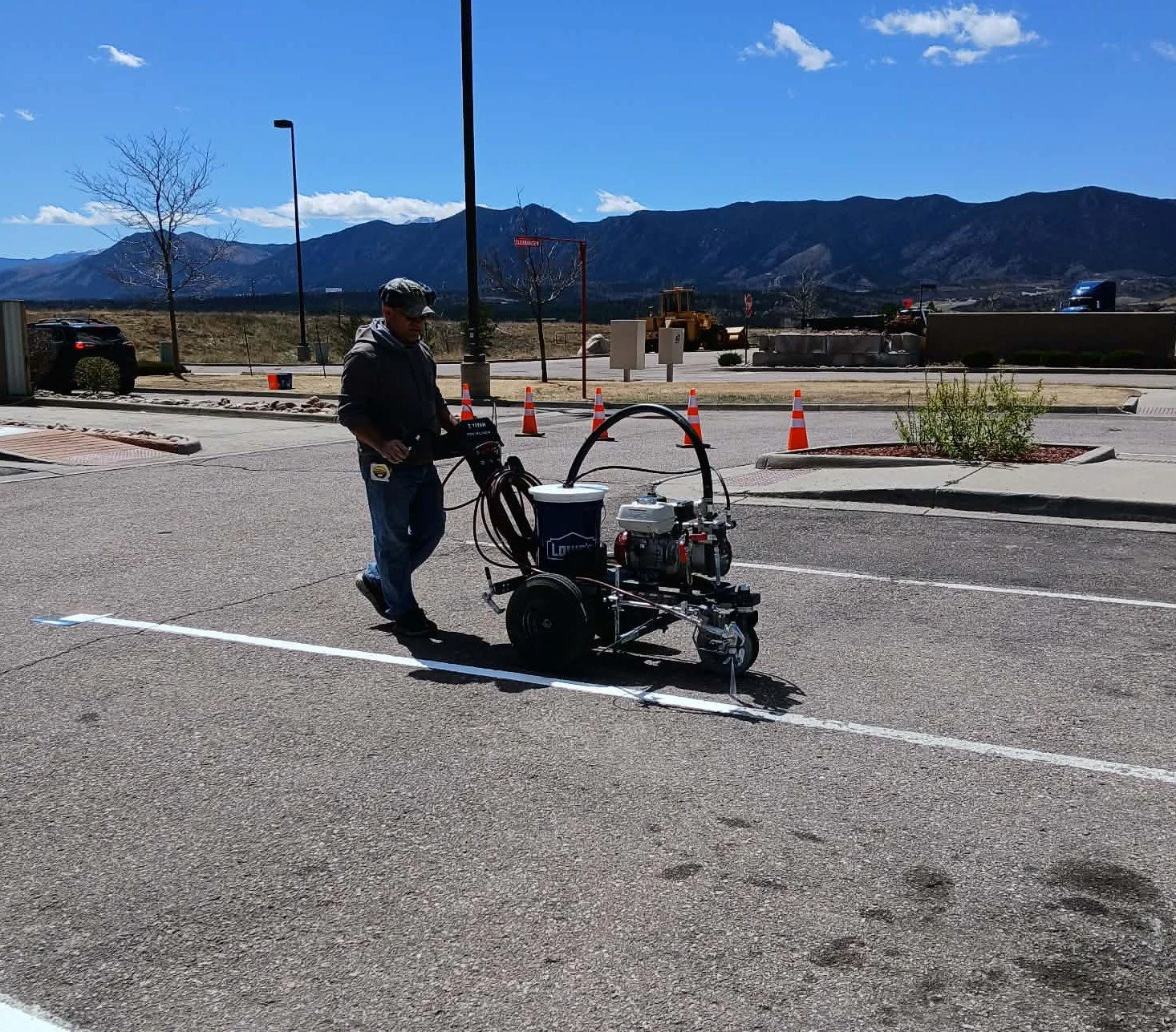 A worker operating a line striping machine in a parking lot with mountains in the background.