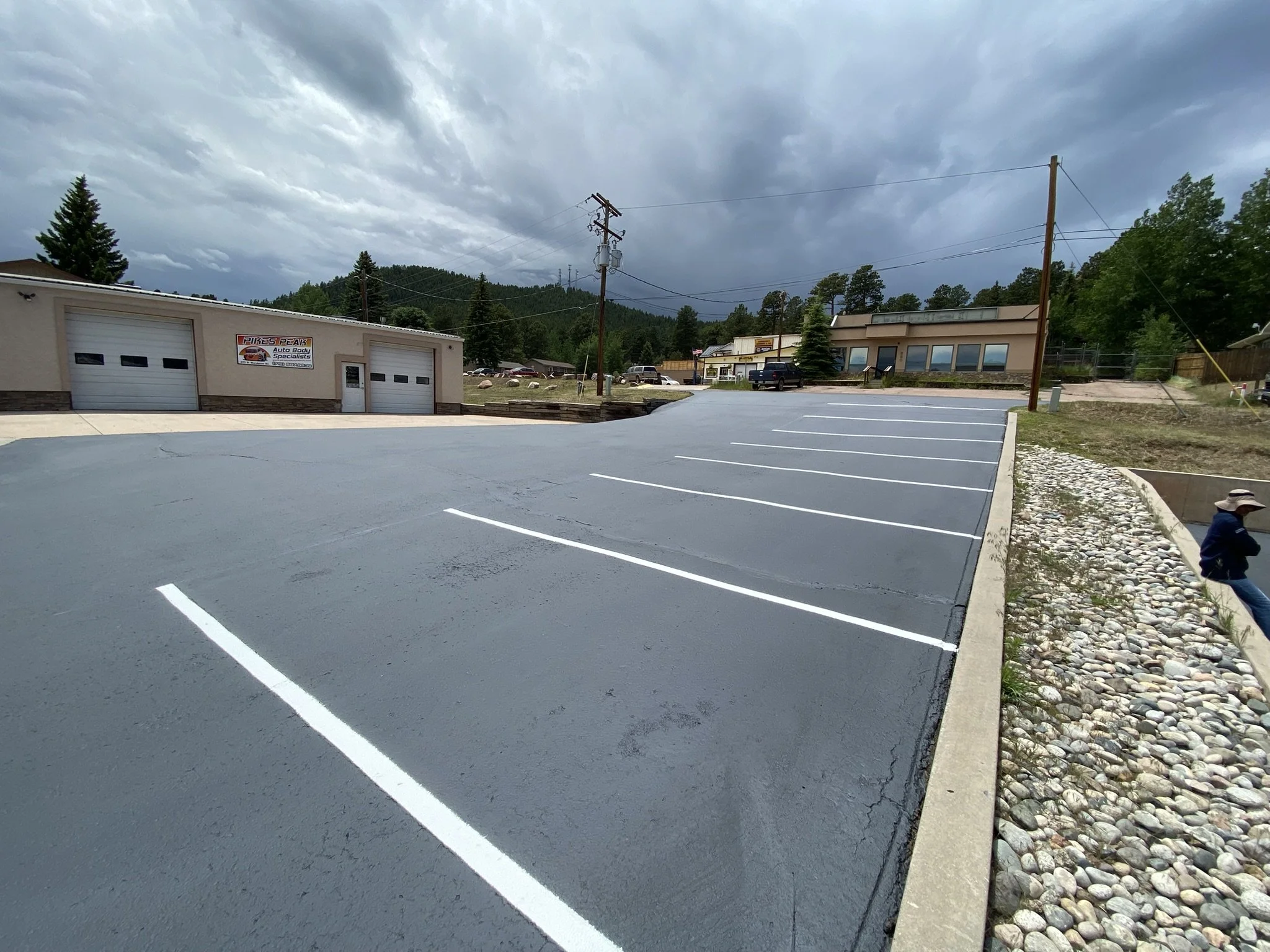 An outdoor parking lot with newly painted white parking space lines, a building with garage doors and a sign for Pikes Peak Auto Body Specialists, several parked cars, power lines, trees, and mountains in the background under a cloudy sky.