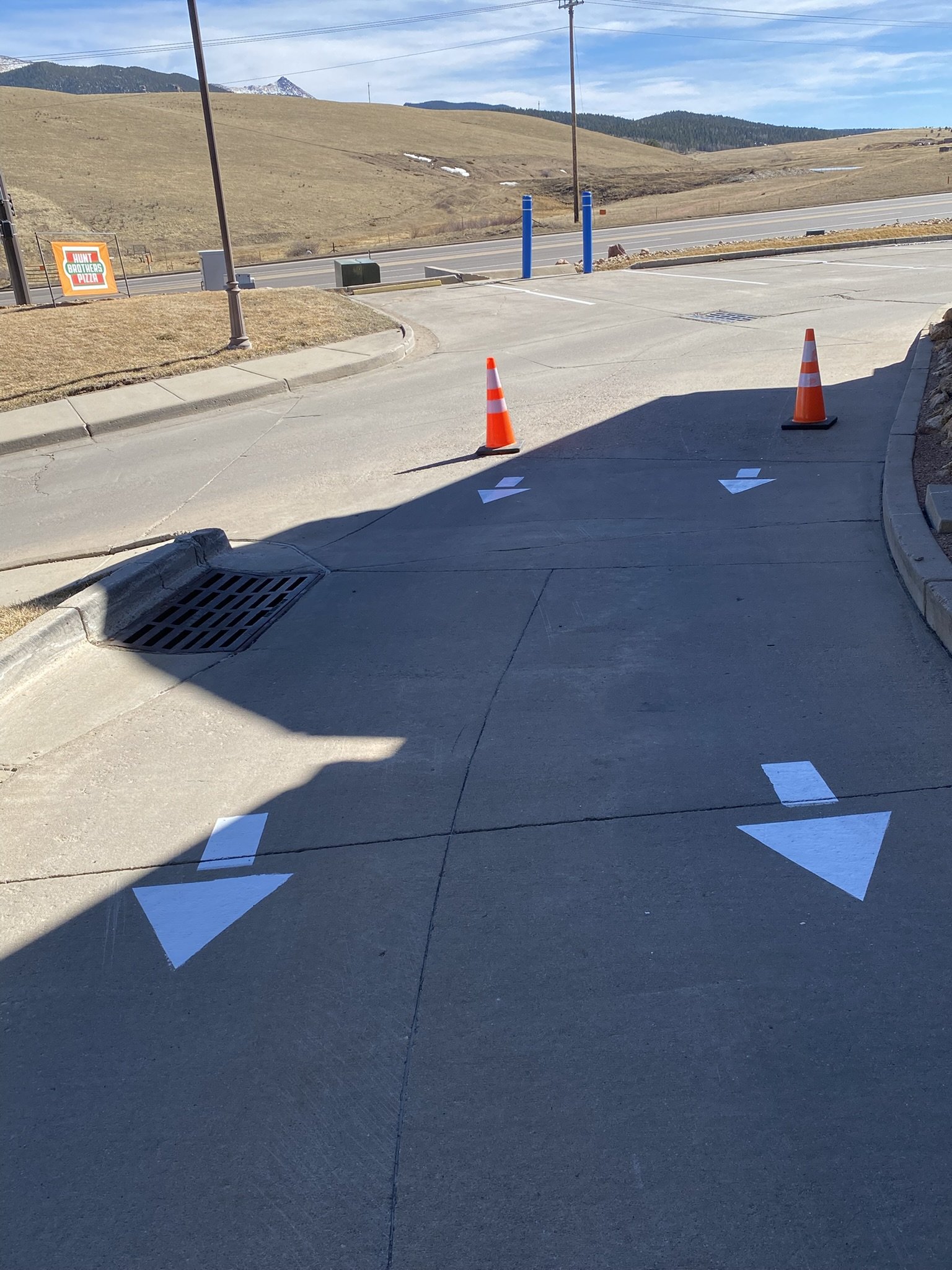 Empty parking lot with orange traffic cones and painted directional arrows on the pavement, with a rural landscape in the background.