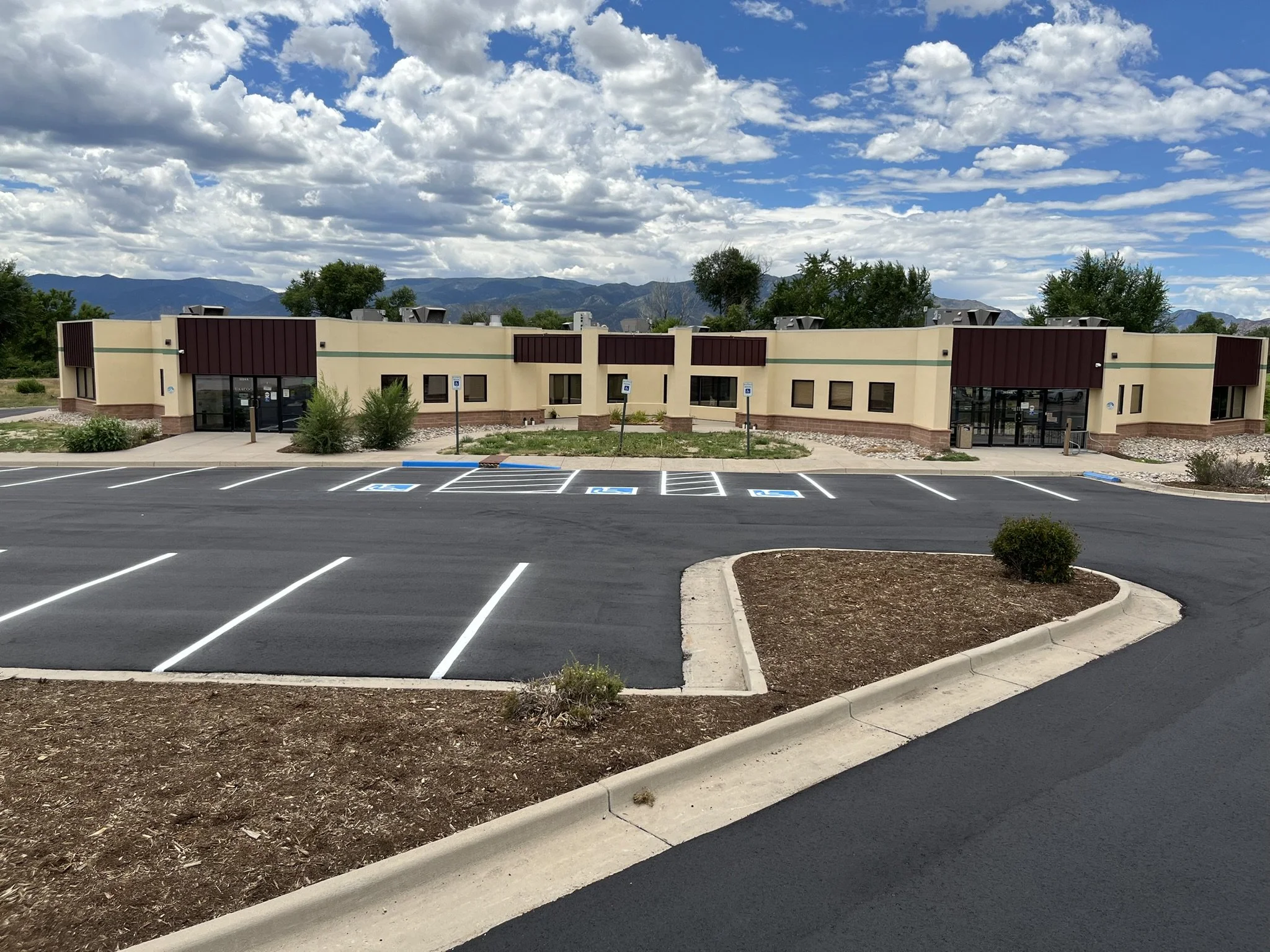 Empty parking lot in front of a modern beige building with large windows, surrounded by landscaped areas with small bushes and trees, under a partly cloudy sky with mountains in the background.