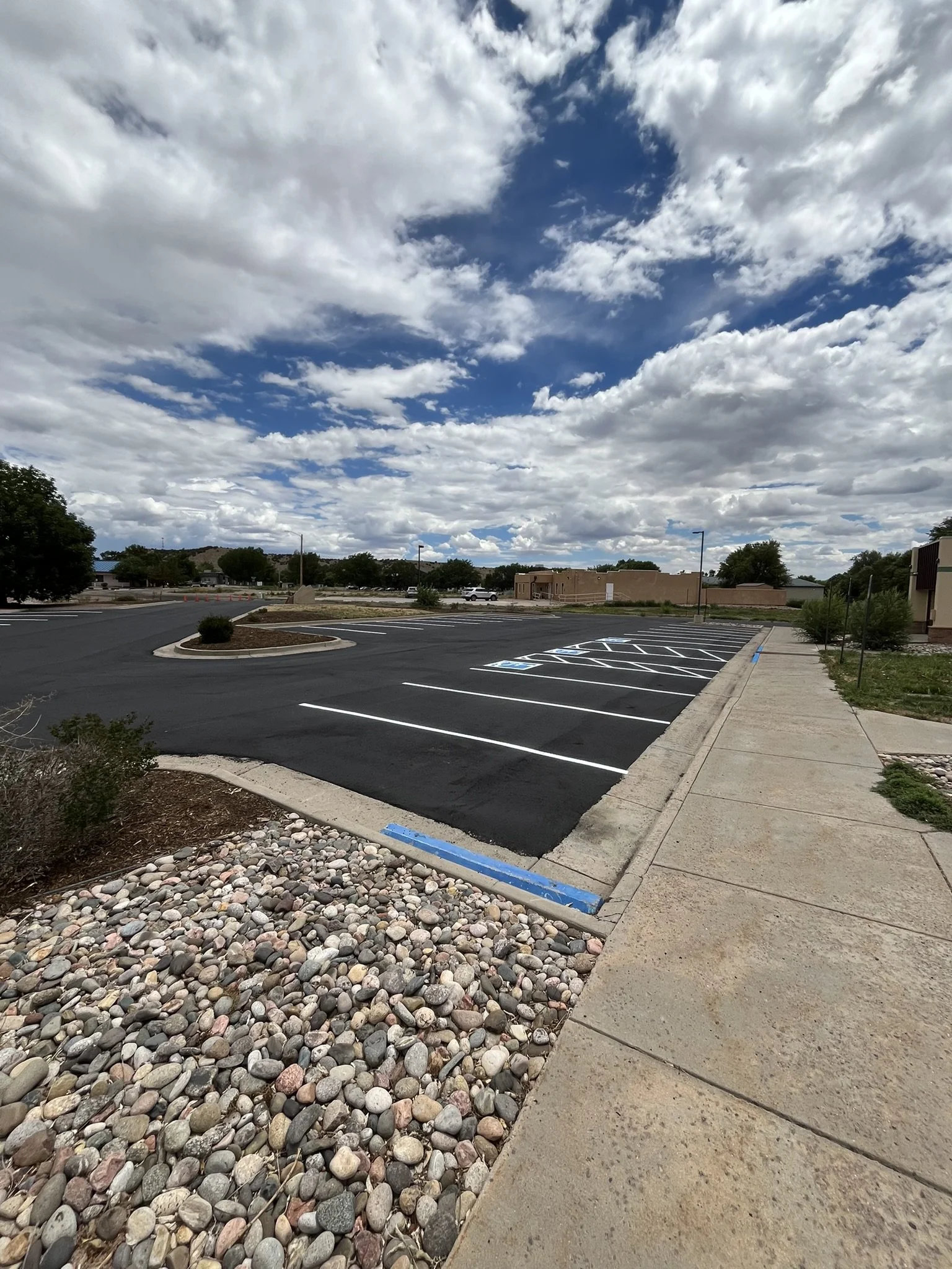 Empty parking lot with newly paved spaces, some marked for handicapped parking, alongside a sidewalk with small shrubs and rocks, under a partly cloudy sky.