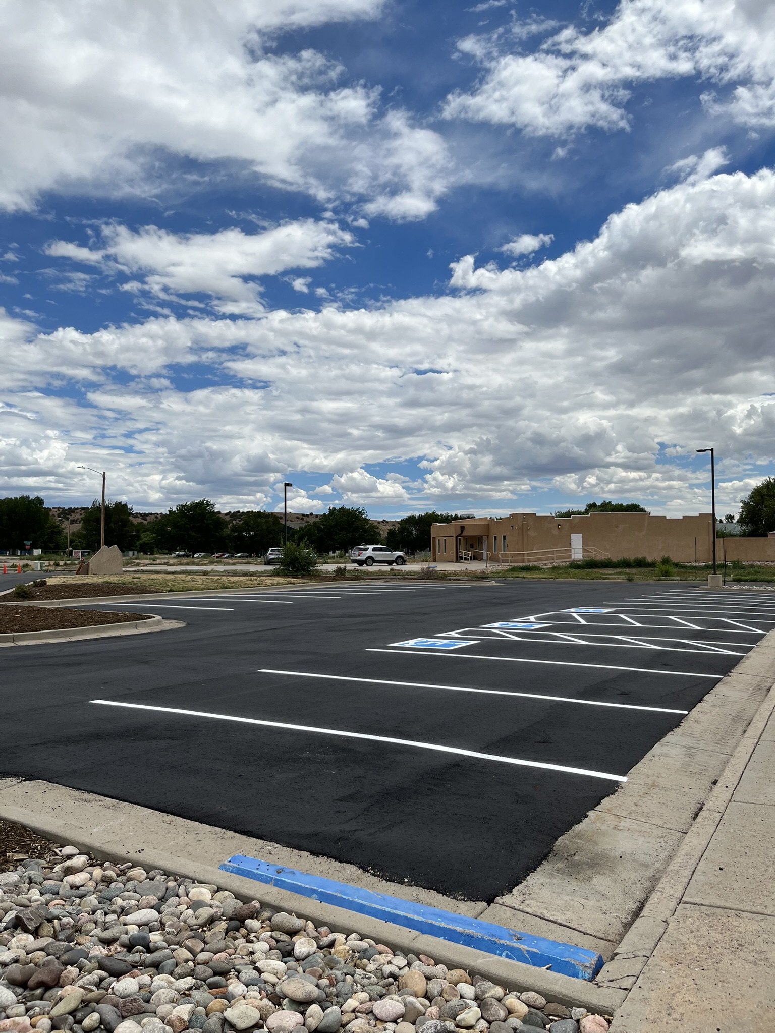 Empty parking lot with freshly paved spaces, small rocks along the curb, and a beige building with a ramp, under a partly cloudy sky.