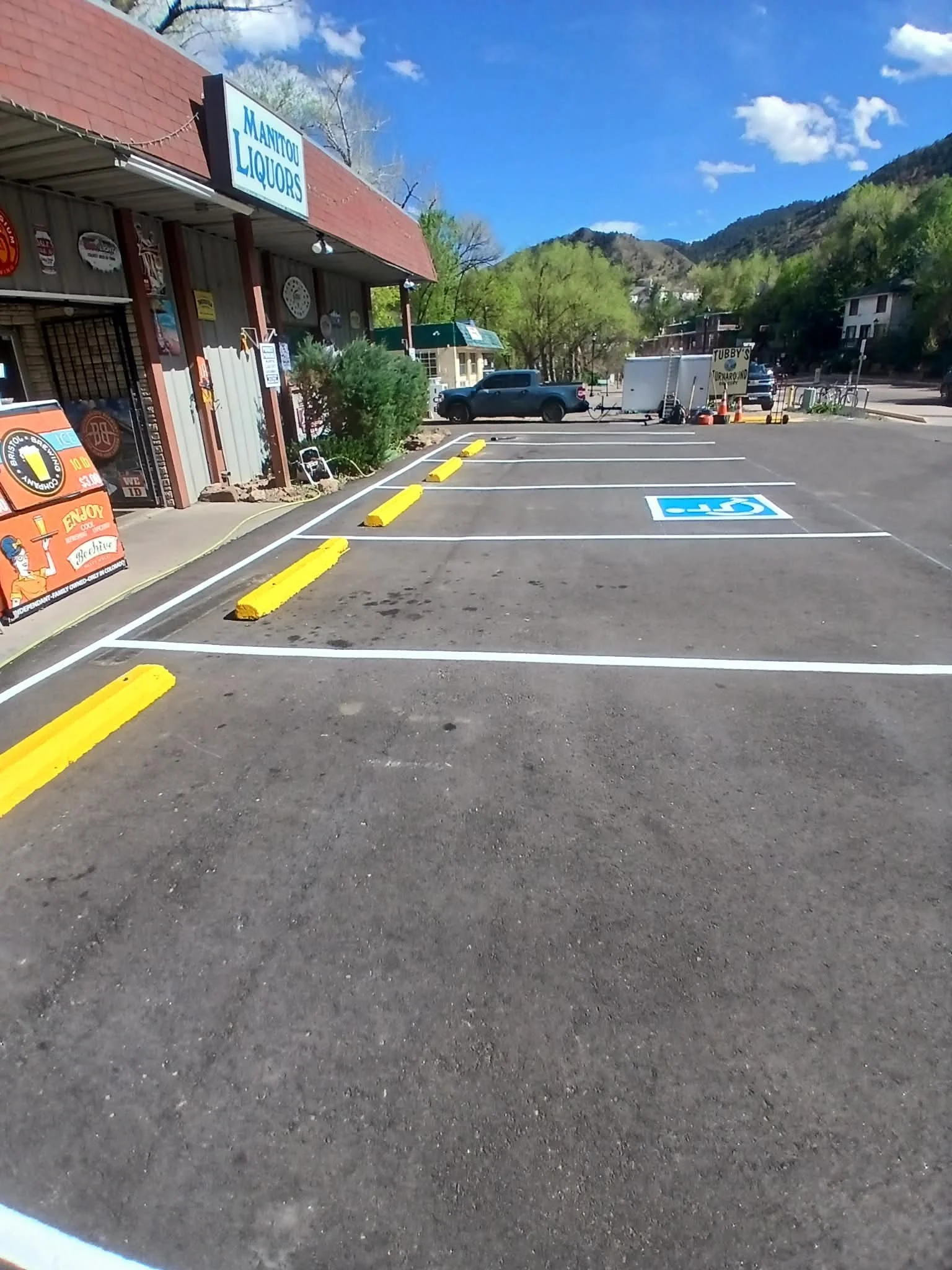 Parking lot with two handicapped spaces marked in blue, empty, with yellow parking blocks. A storefront named 'Manitou Liquors' is on the left side. There is a small trailer and a black pickup truck in the background, with green trees and mountain hi